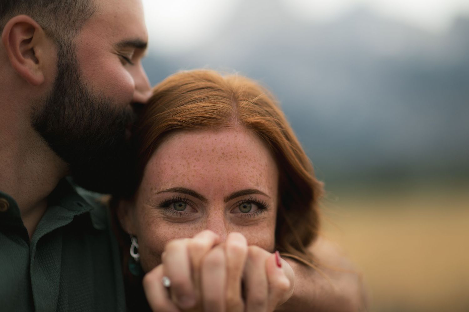 A close-up portrait of a man embracing a woman with a mountainous landscape in the background.