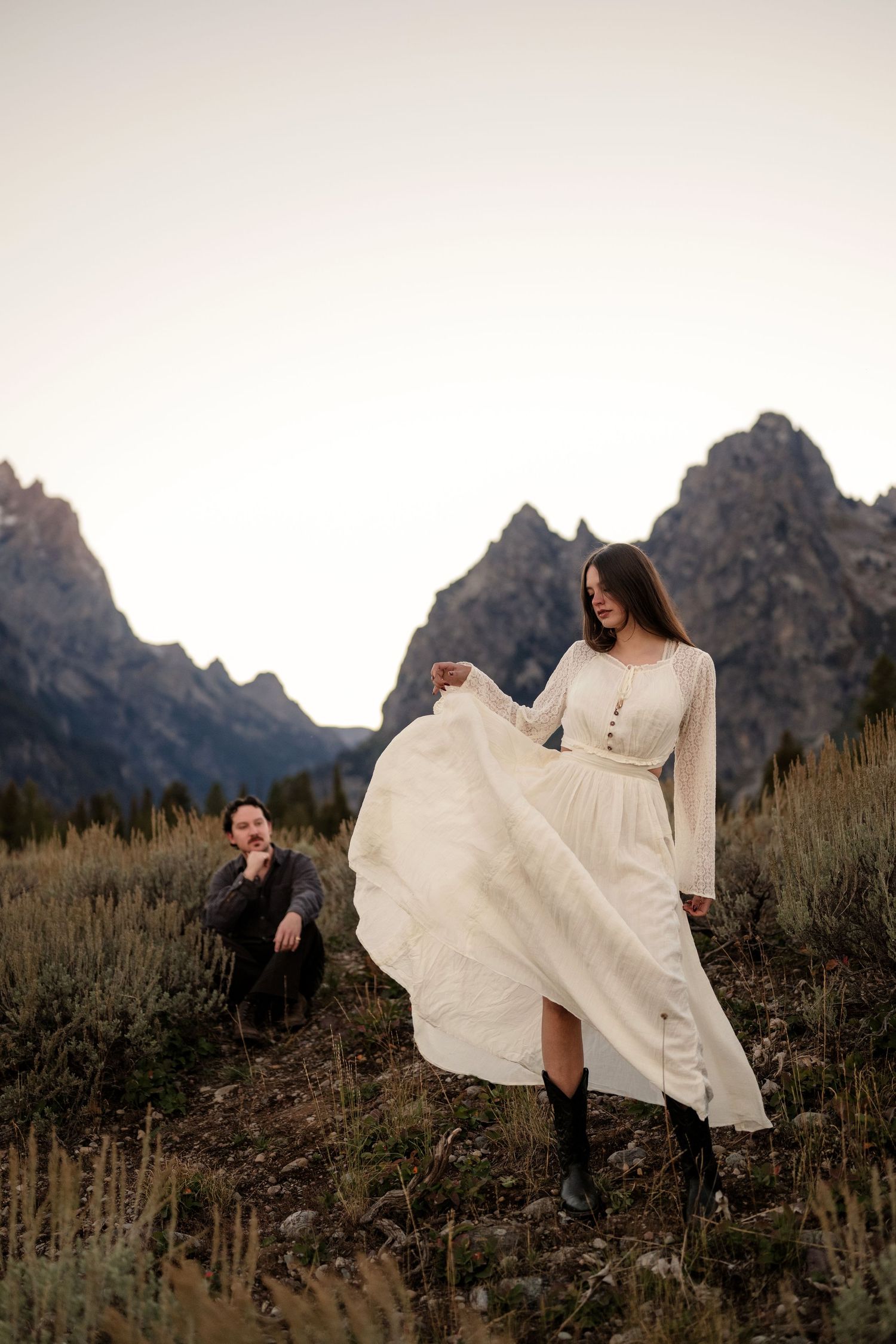 Bride in white dress and cowboy boots twirls skirt with groom seated behind her, Grand Teton mountains at sunset.