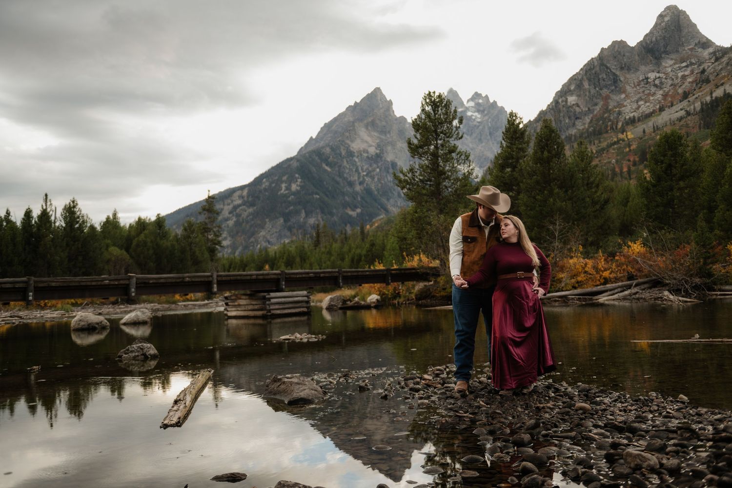 Couple embraces near a rocky river with Grand Teton mountains and autumn trees in the background on a cloudy day.