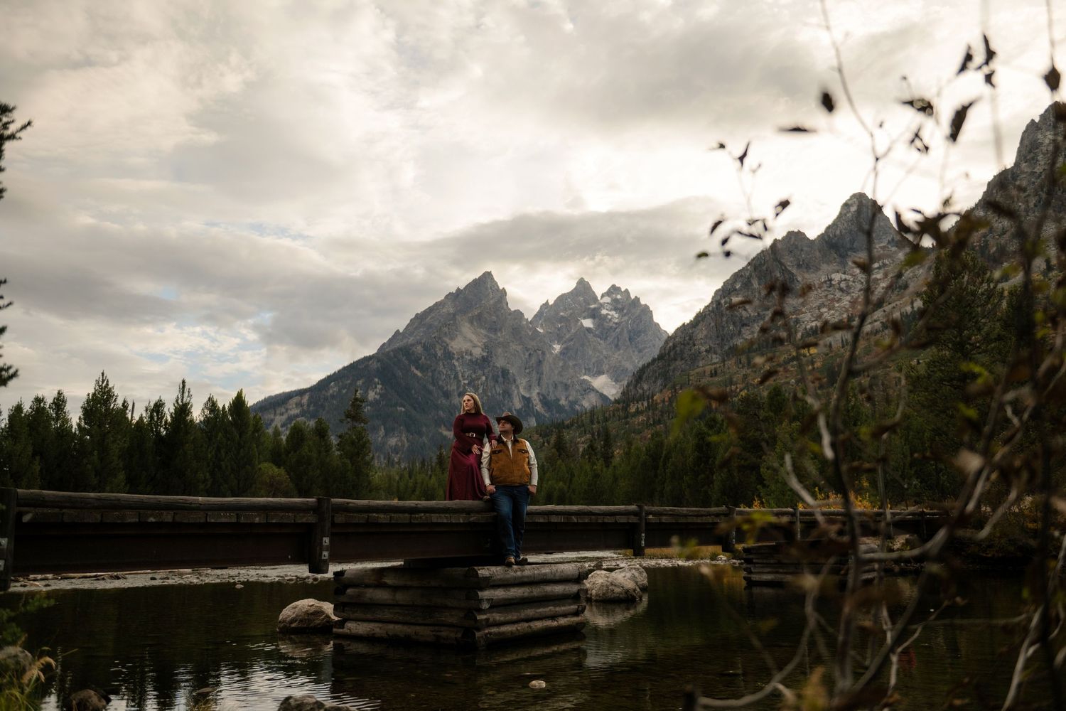 A couple stands on a wooden bridge over a lake, with snow-capped mountains in the background.
