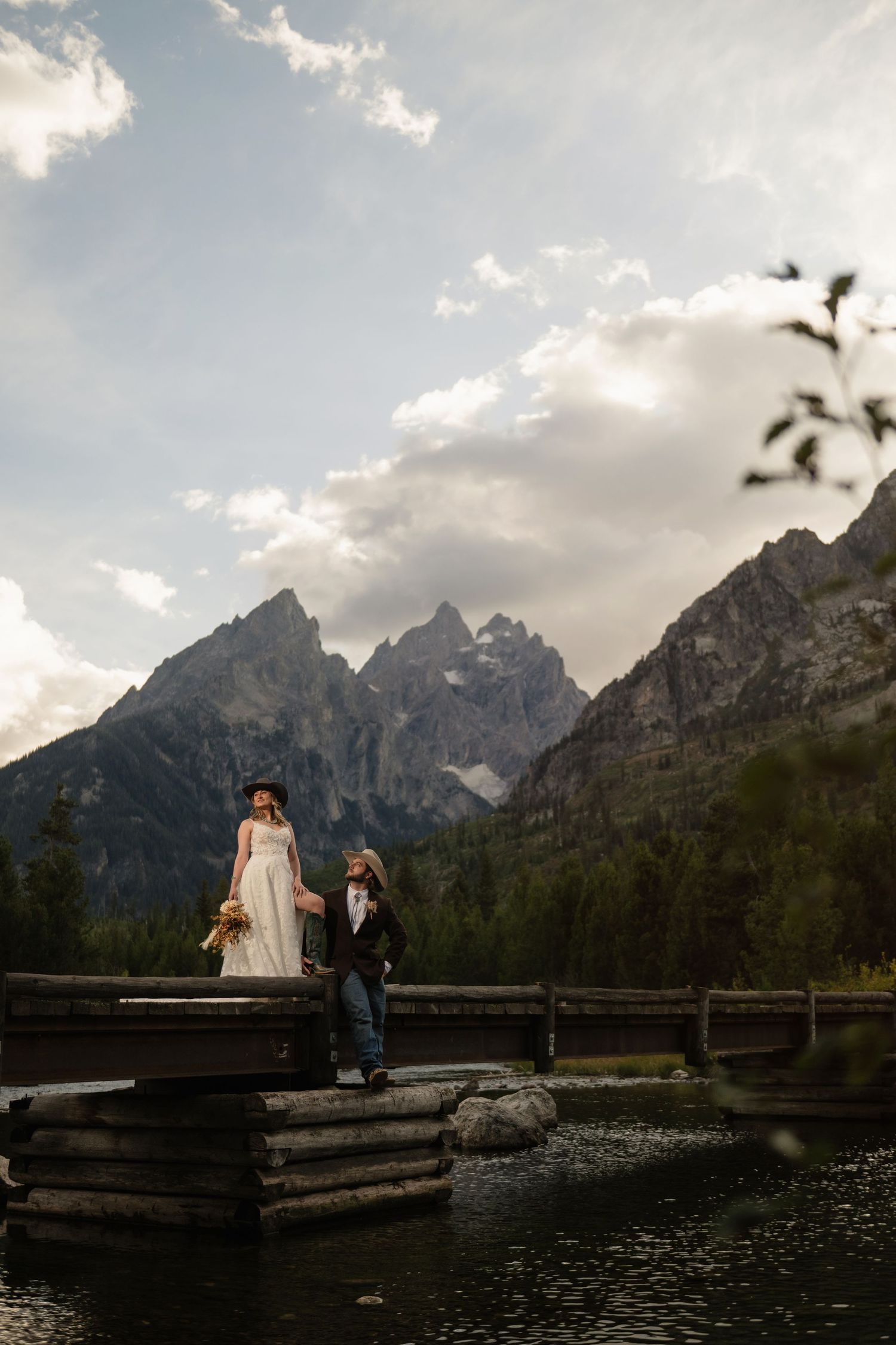 Bride and groom stand on a rustic wooden bridge with Grand Teton mountains and a calm lake in the background.