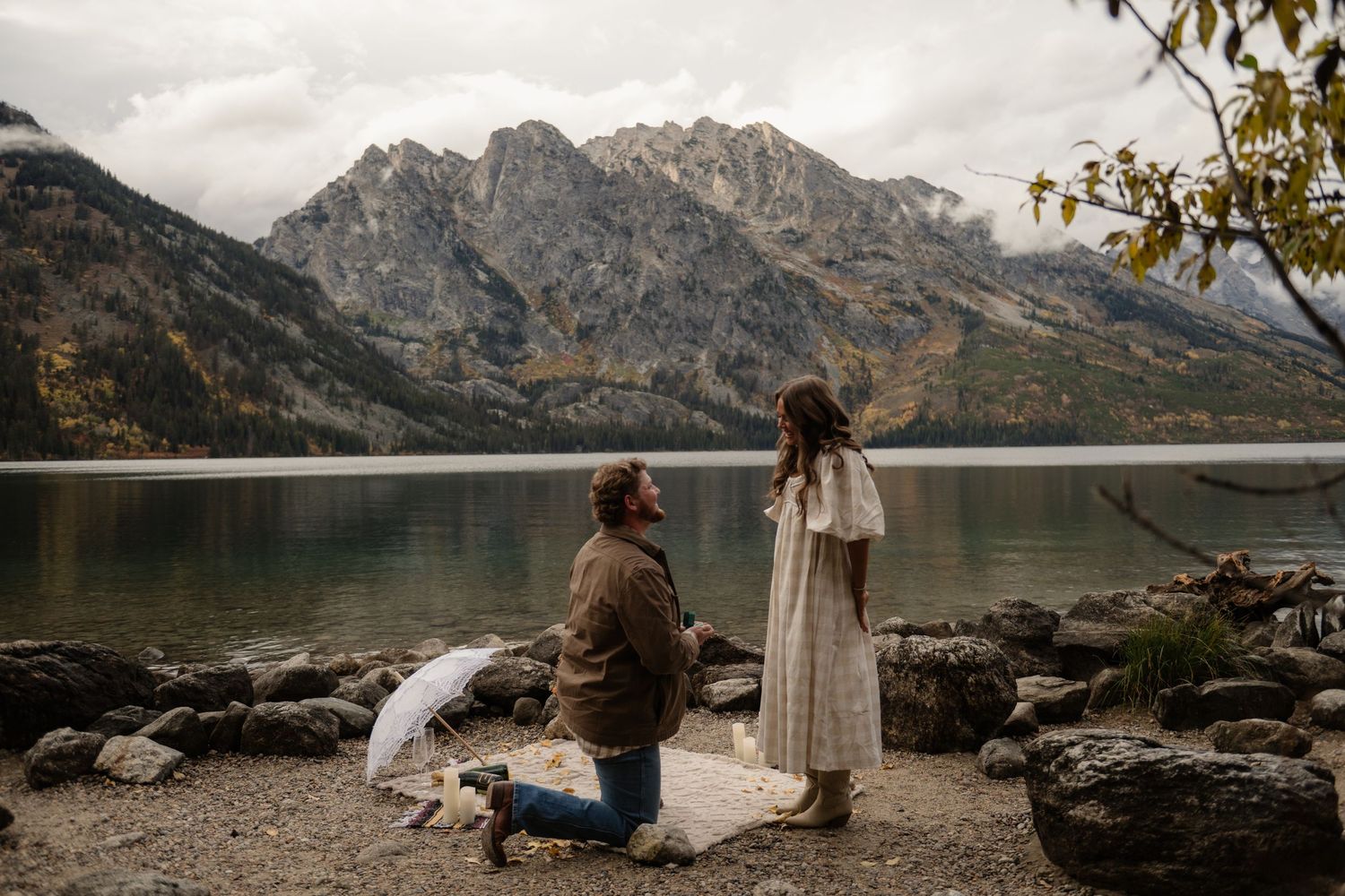 Man proposes on one knee by a mountain lake with stunning fall foliage and rocky shoreline backdrop.