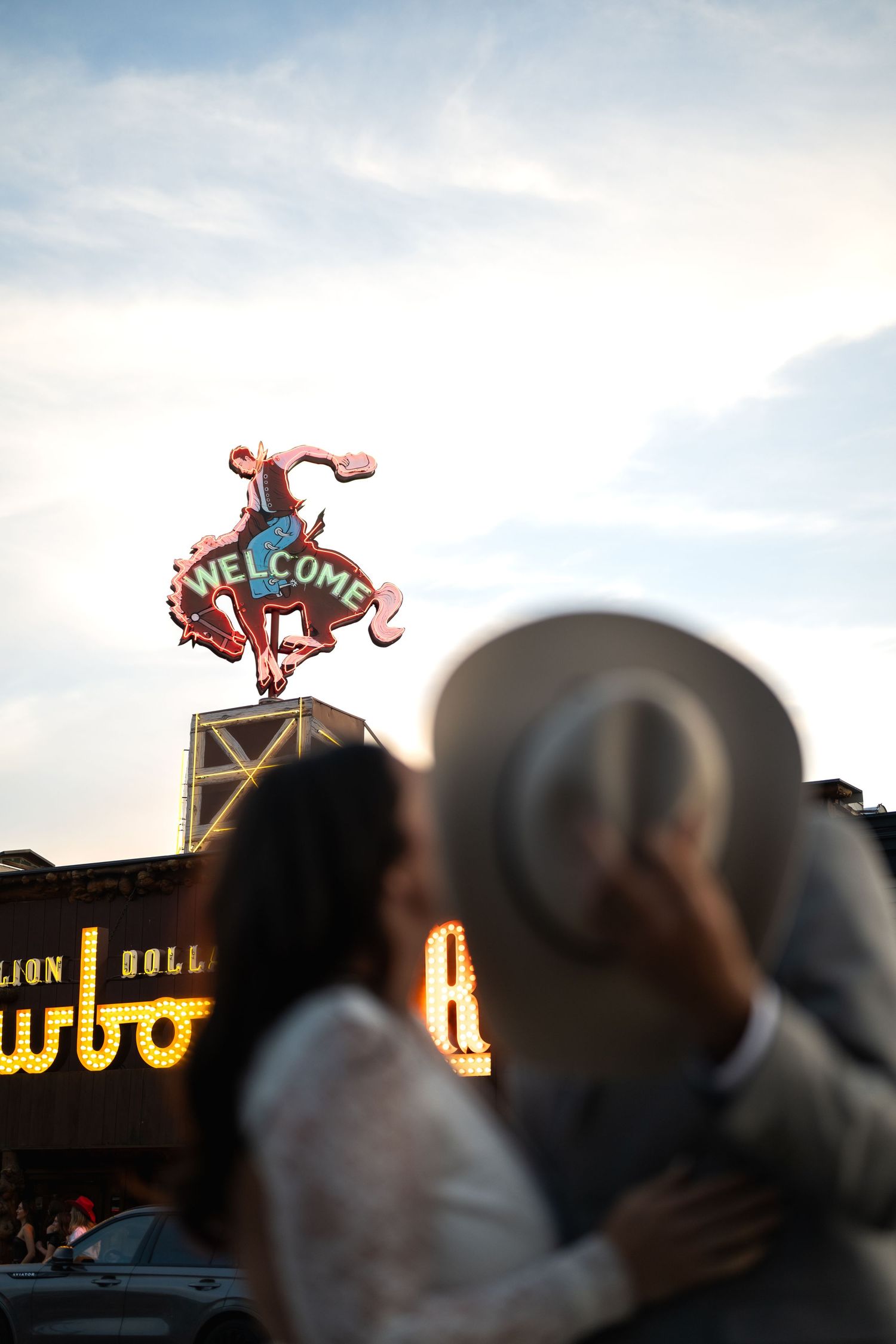 Couple embracing near a glowing Welcome neon cowboy sign at dusk in a vibrant city setting.