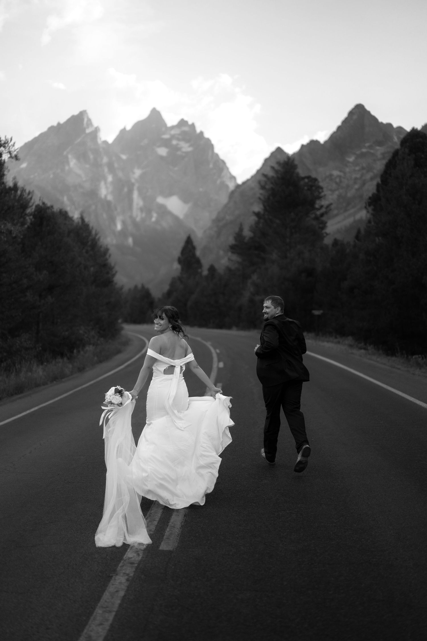 A couple walks down a road in a mountainous landscape, with the woman wearing a long white dress and the man wearing a suit.