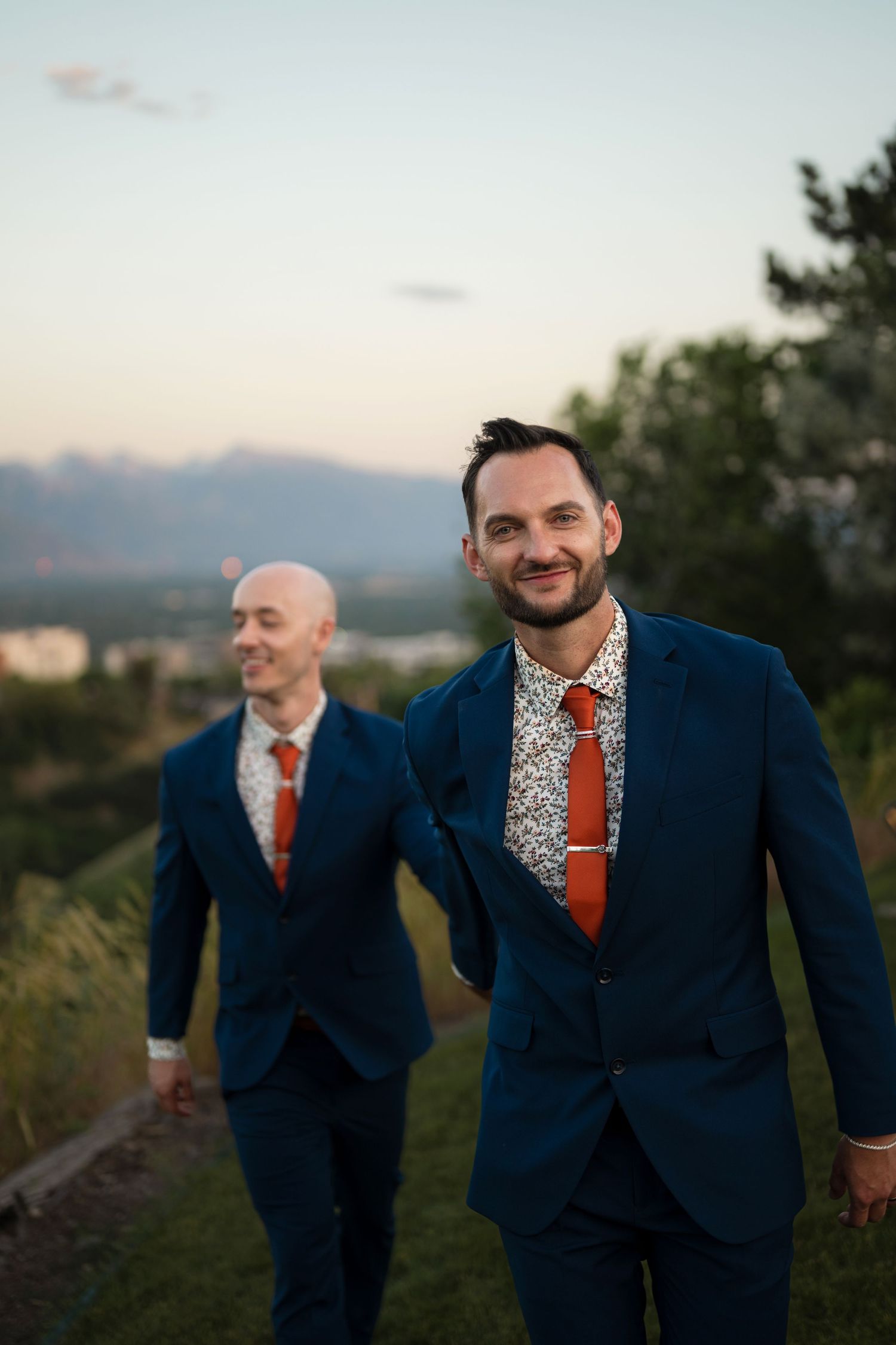 Two grooms in matching navy suits and orange ties walk hand in hand outdoors at sunset.