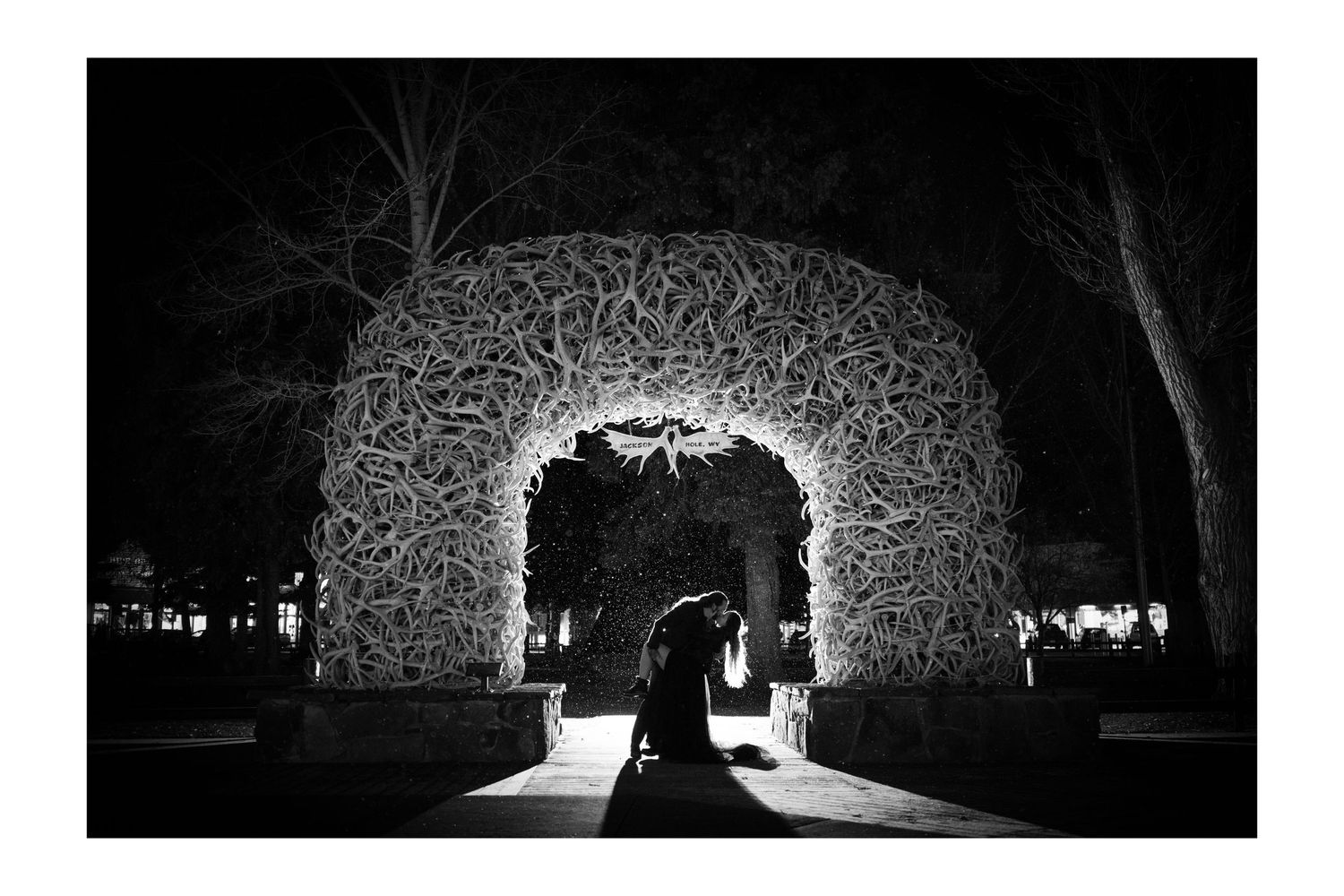 Black and white photo of a couple silhouetted under an illuminated antler arch at night in Jackson Wyoming