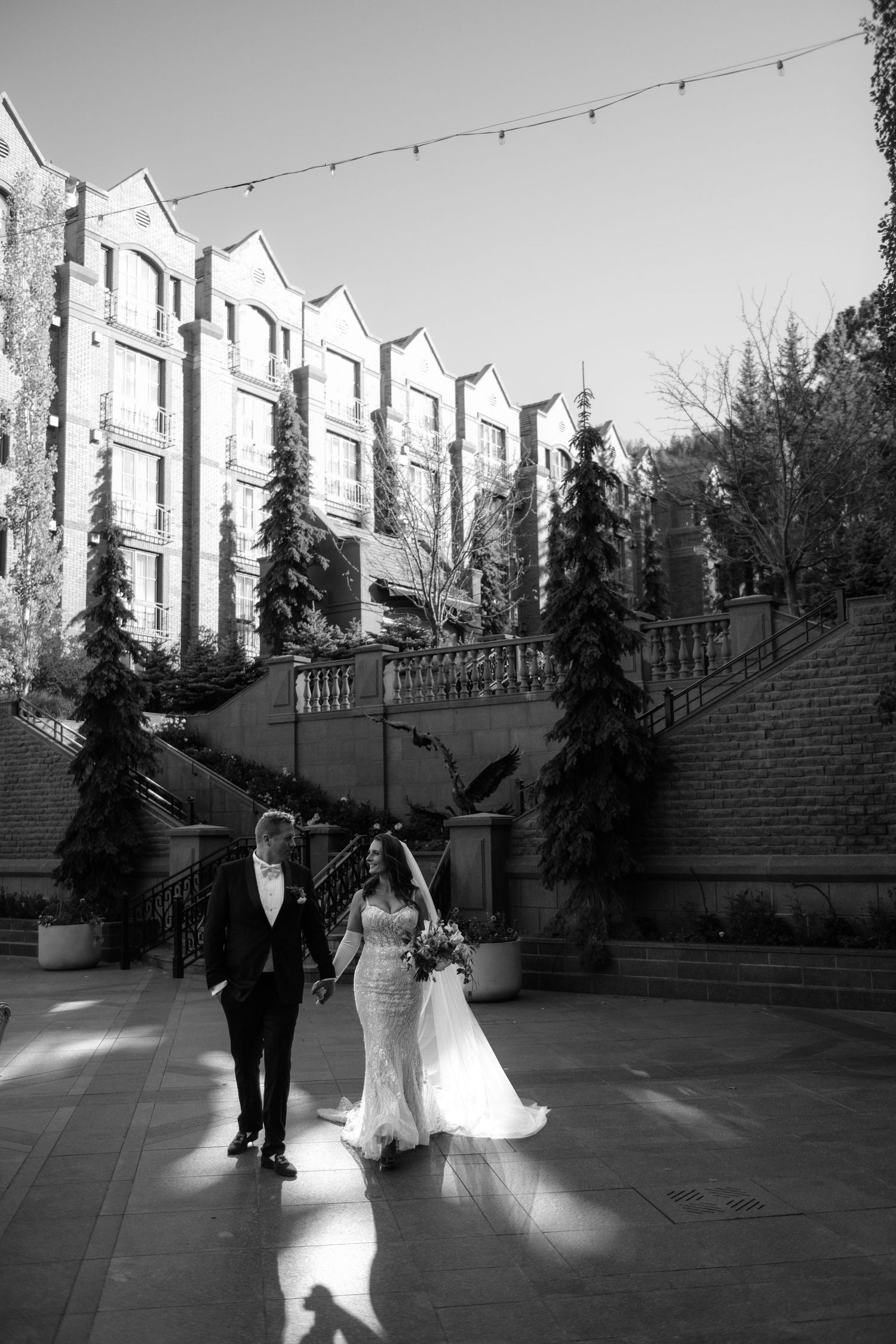 A black and white image shows a couple walking together in an outdoor setting with string lights overhead.