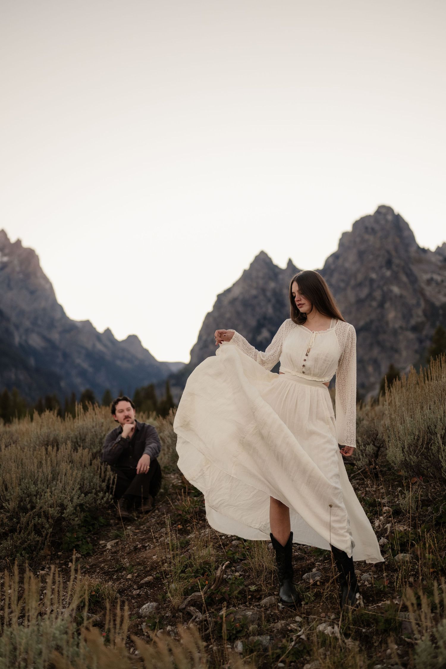 Couple in mountain meadow, woman in white boho dress with cowboy boots twirls skirt, man in dark jacket, Grand Tetons backdrop.