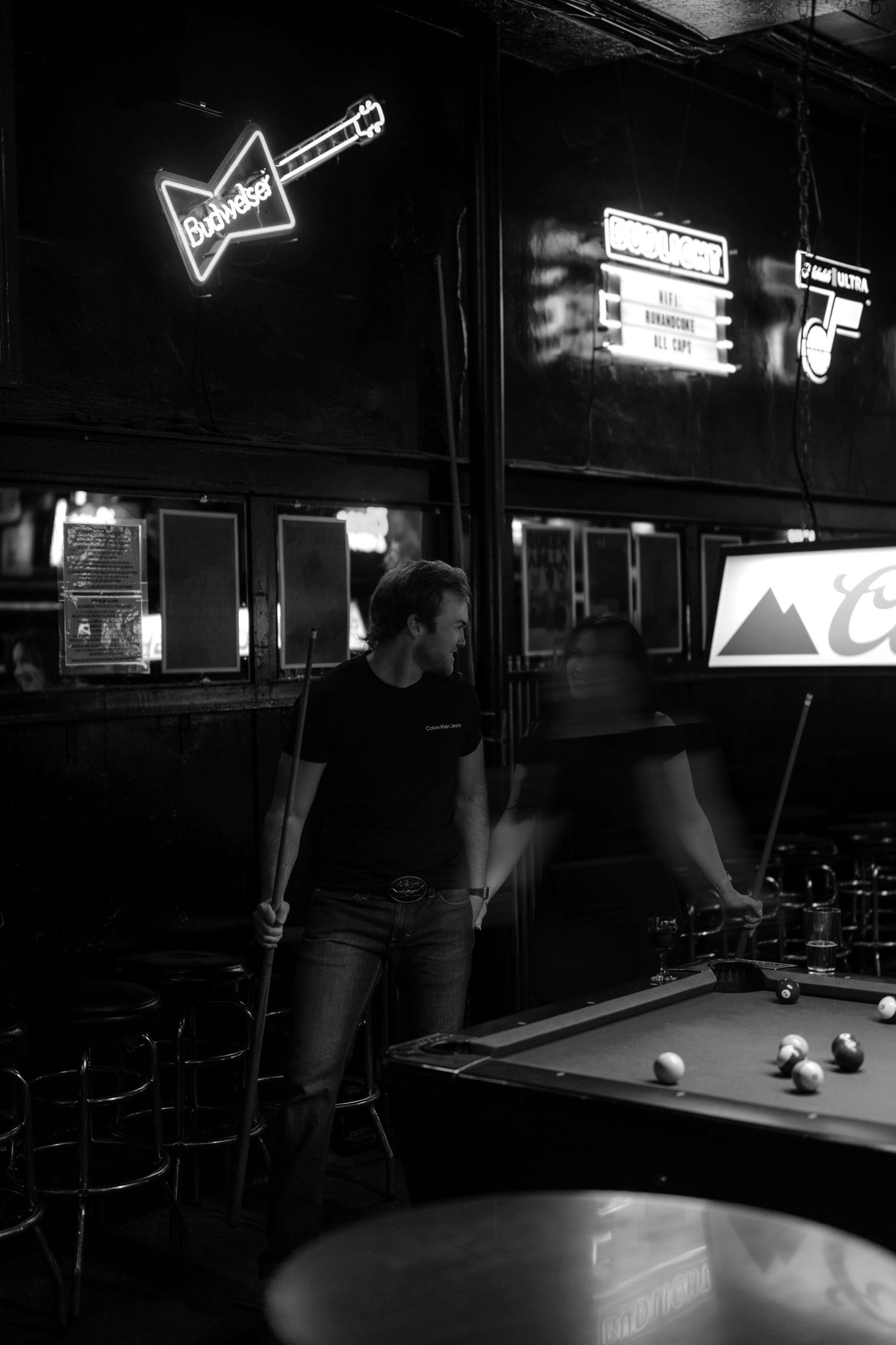 Black and white photo of a woman playing pool in a dimly lit bar with neon Budweiser sign.