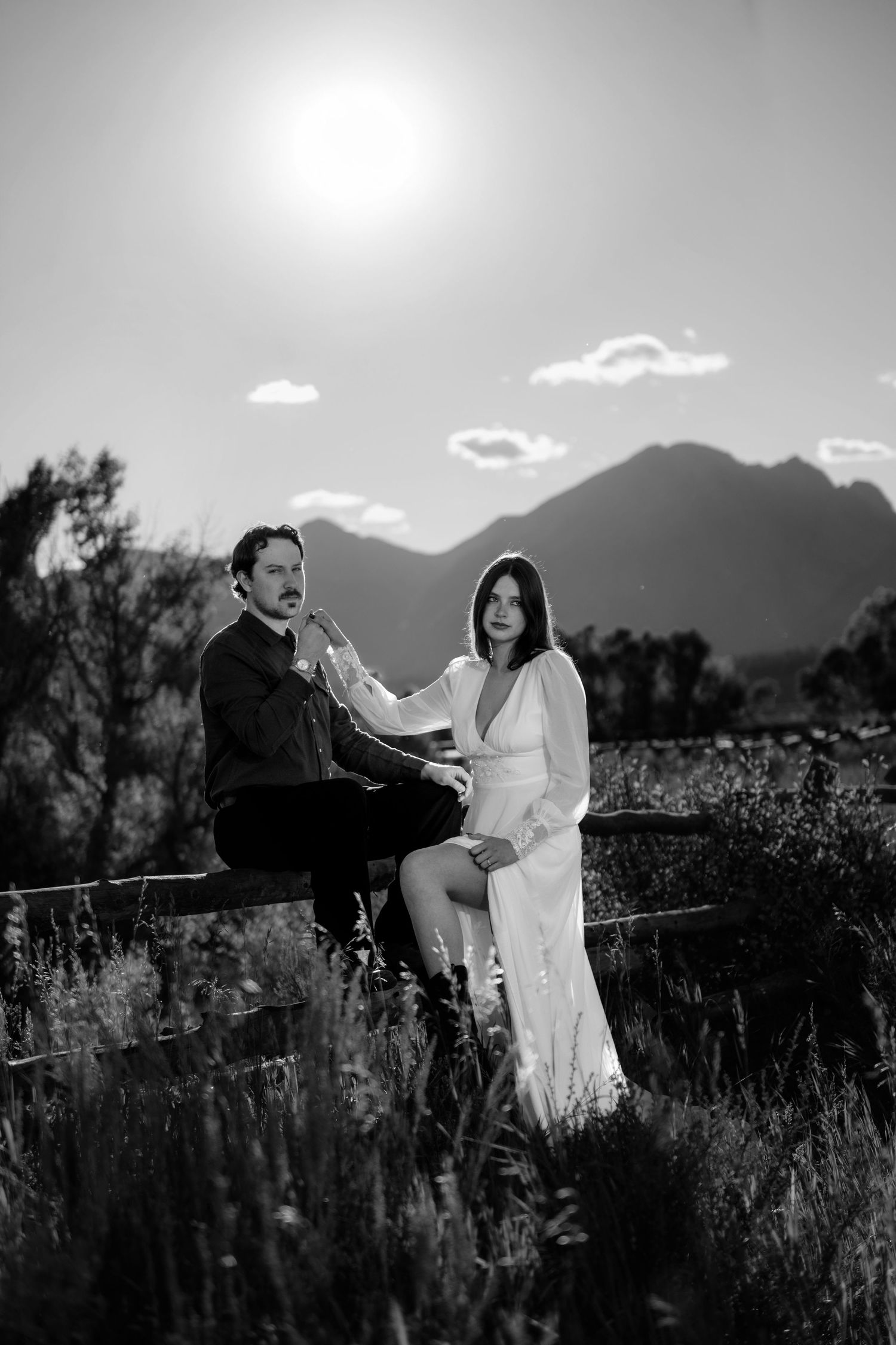 Black and white photo of a couple in wedding attire sitting on a fence with mountains and bright sun in the background.