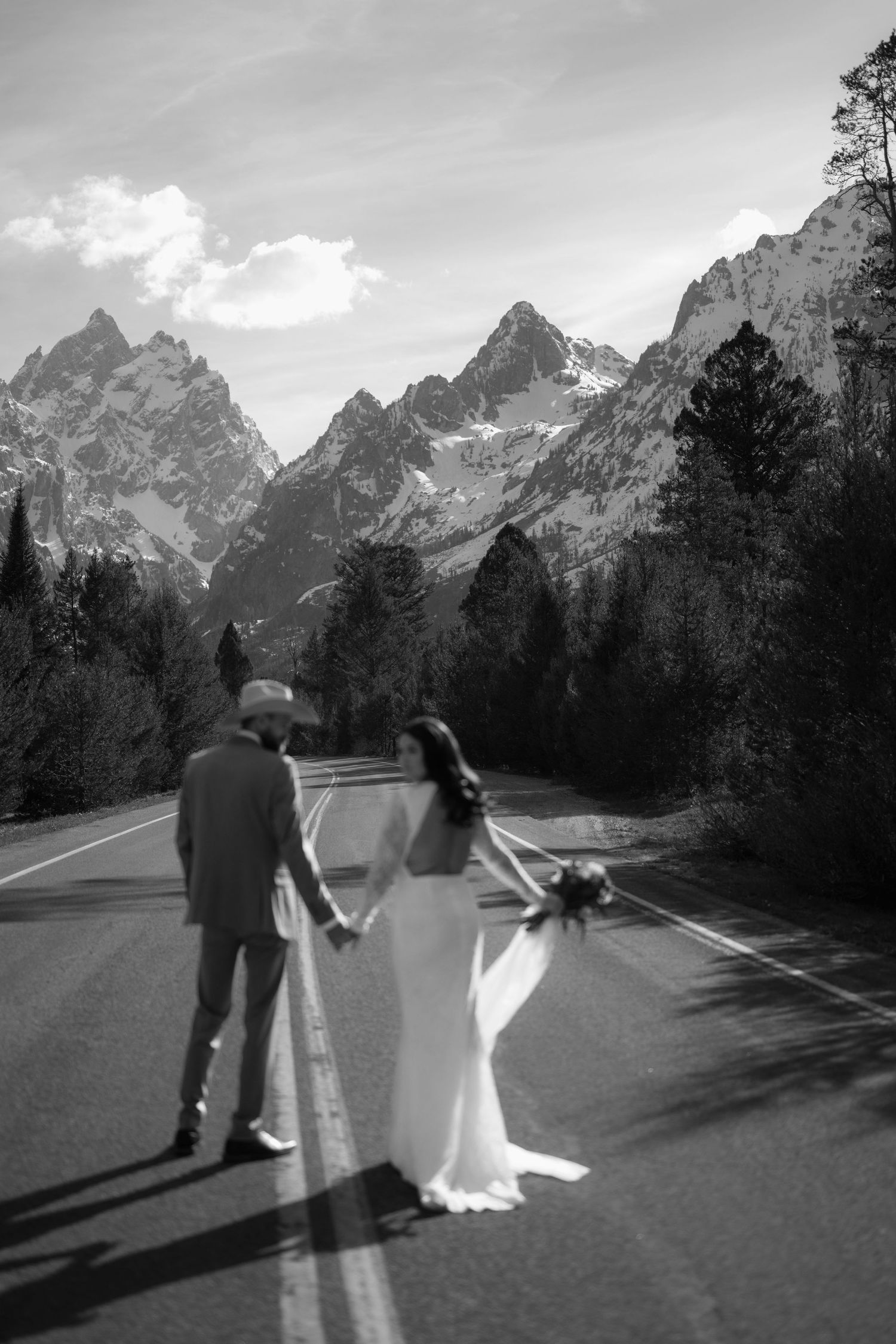Black and white photo of a bride and groom holding hands on a mountain road with Grand Teton peaks in the background.