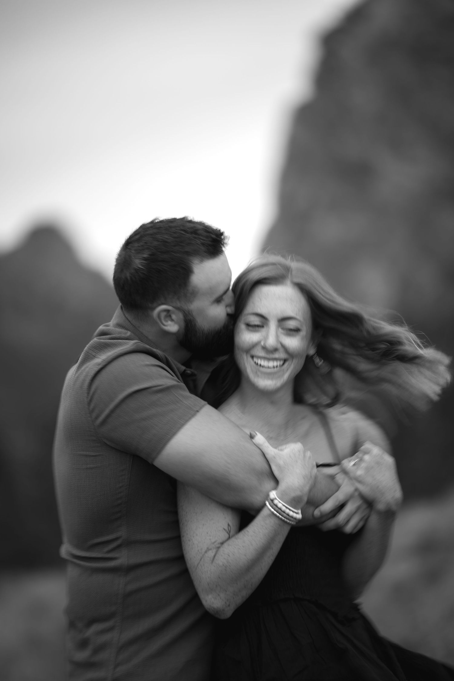 Black and white photo of a laughing woman being embraced and kissed on the cheek by a man outdoors.