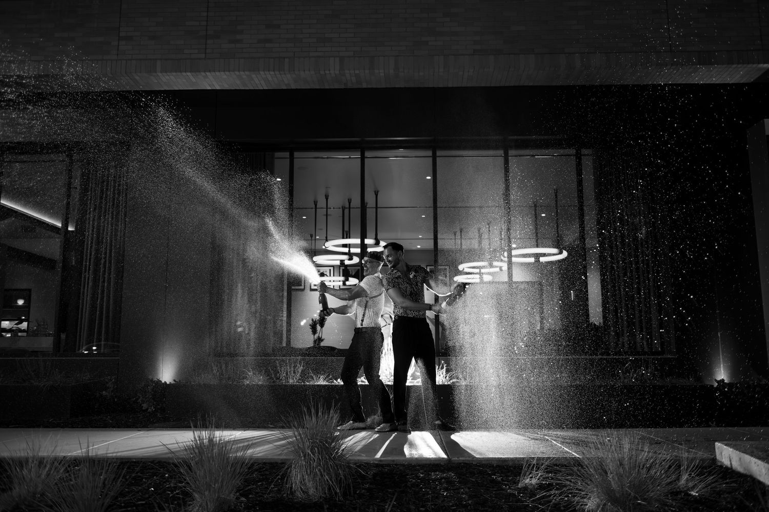 Black and white photo of two people celebrating outdoors at night with champagne spray near a modern building.