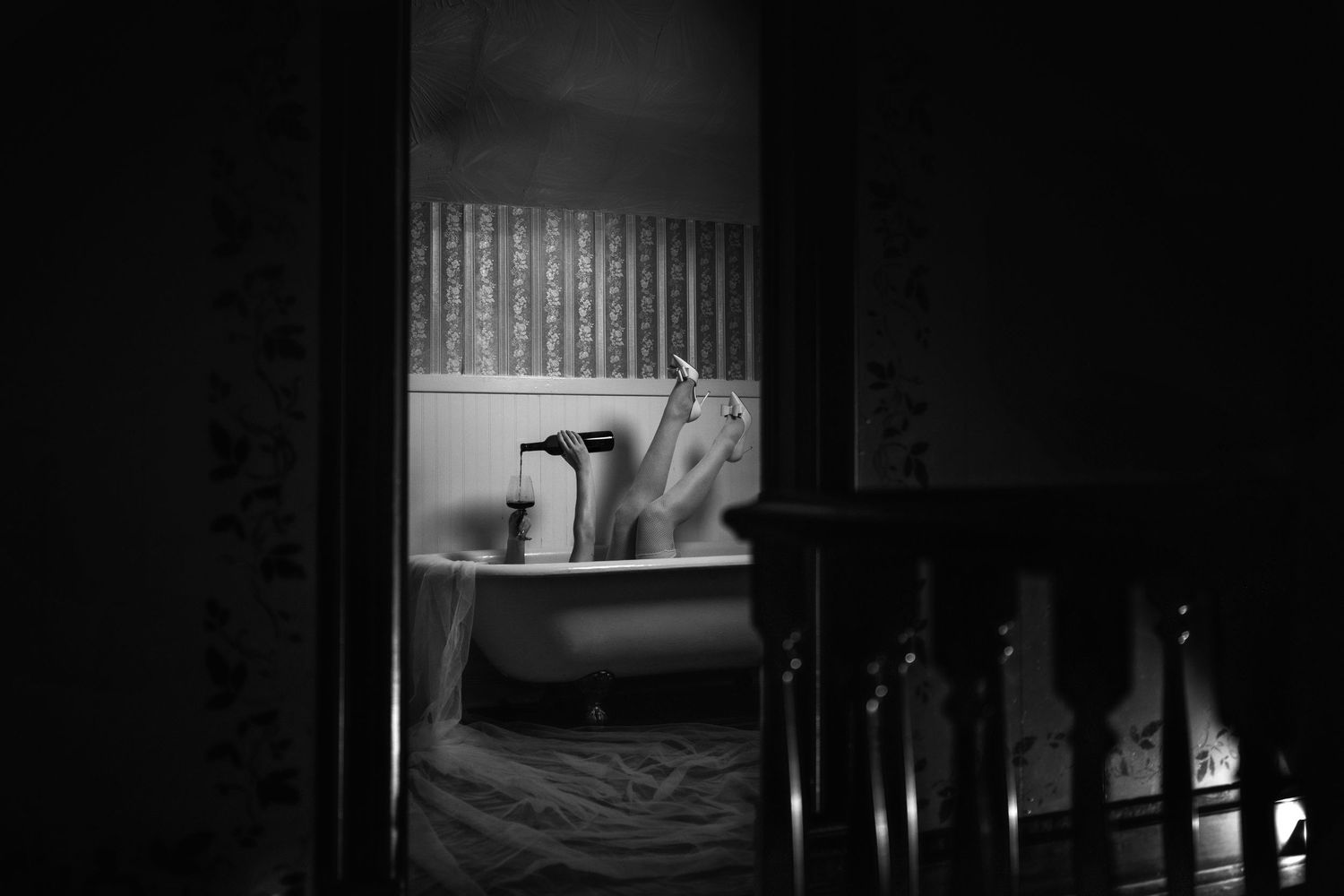 Black and white photo of a person relaxing in a clawfoot bathtub, viewed through a doorway in a dimly lit room.