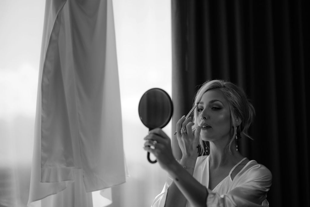 Bride applying makeup while looking in handheld mirror in black and white photo.