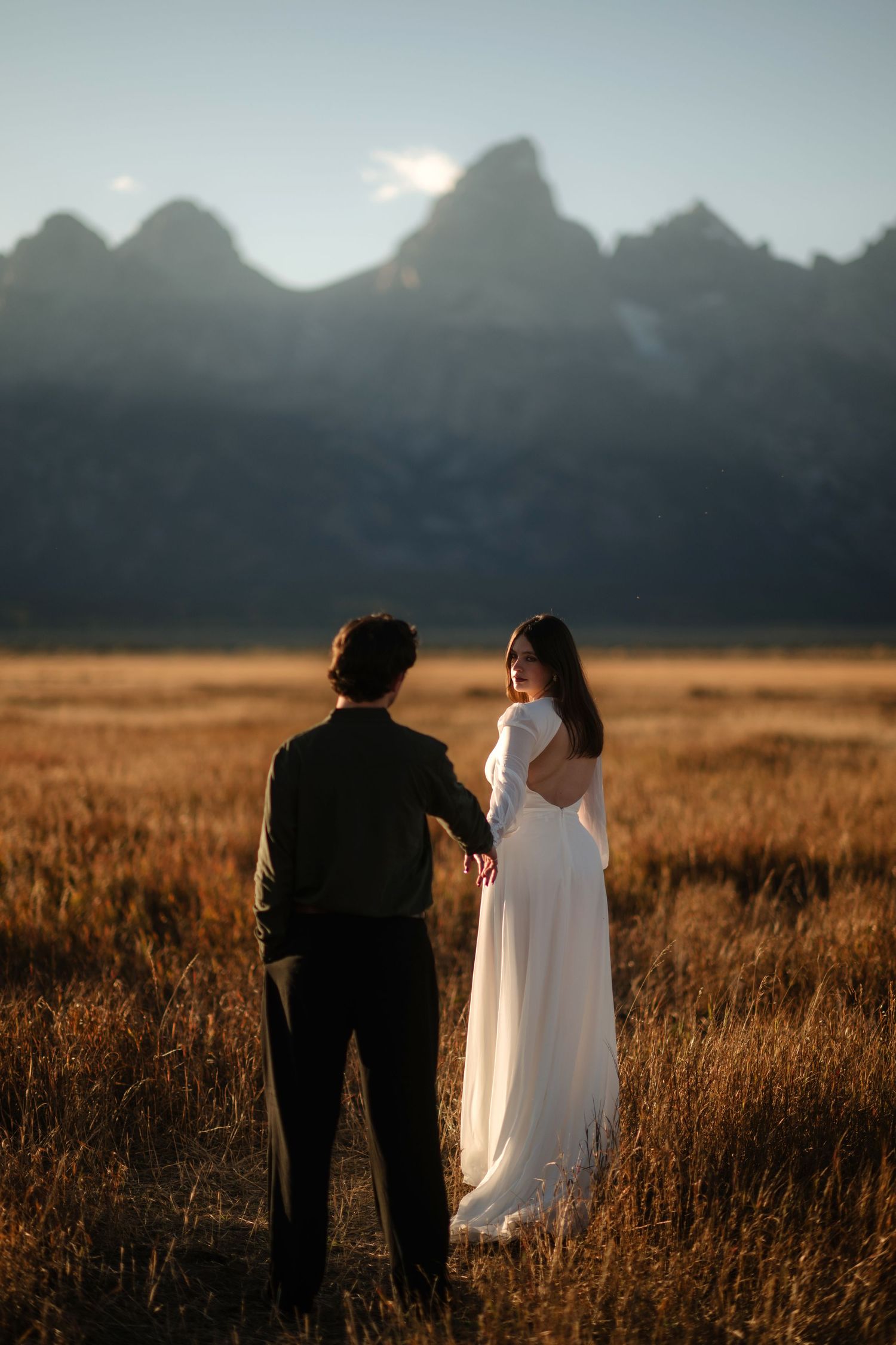 Bride and groom hold hands in golden field with Grand Teton mountains at sunset.