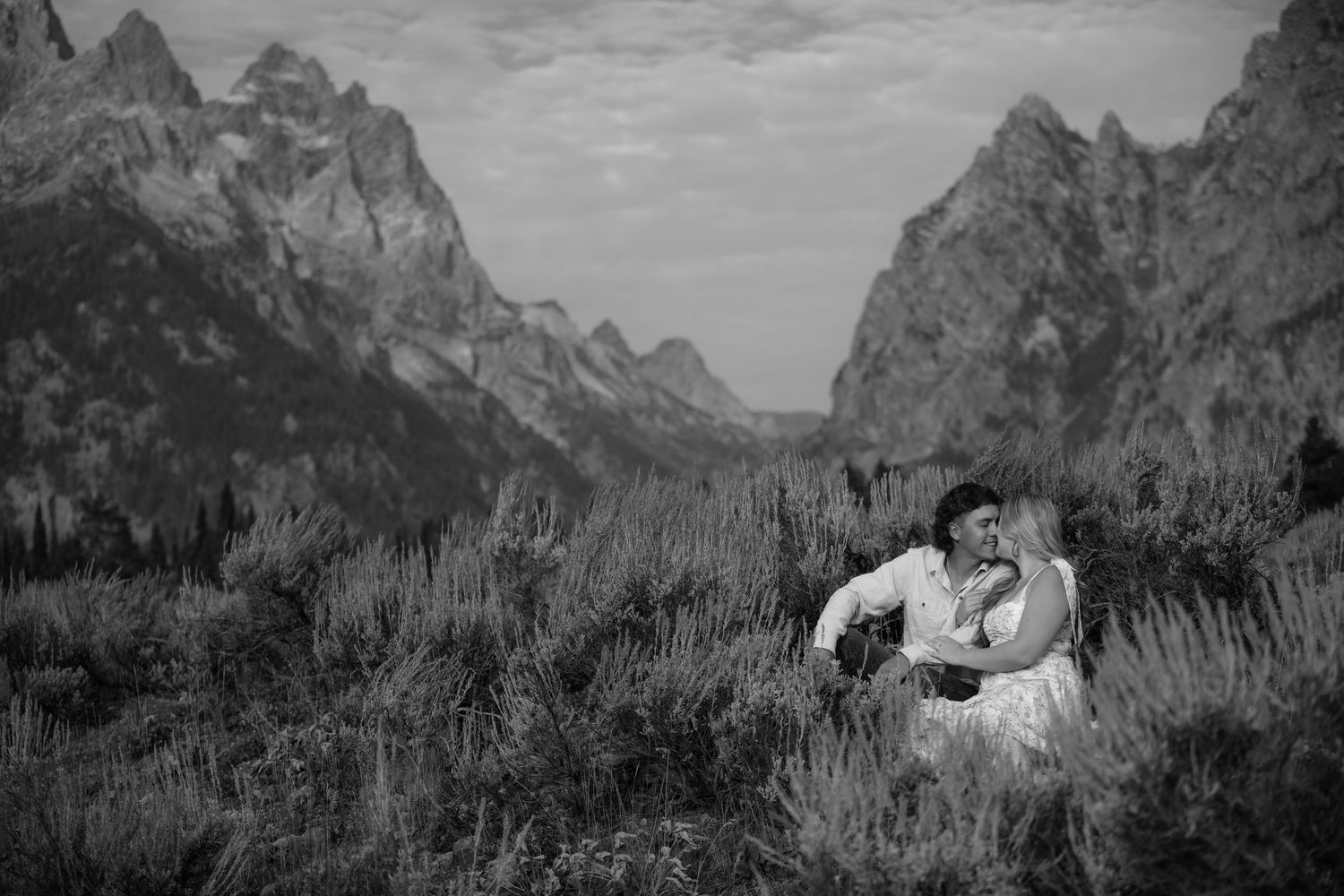Black and white photo of a couple embracing in a field with the Grand Teton mountains in the background.