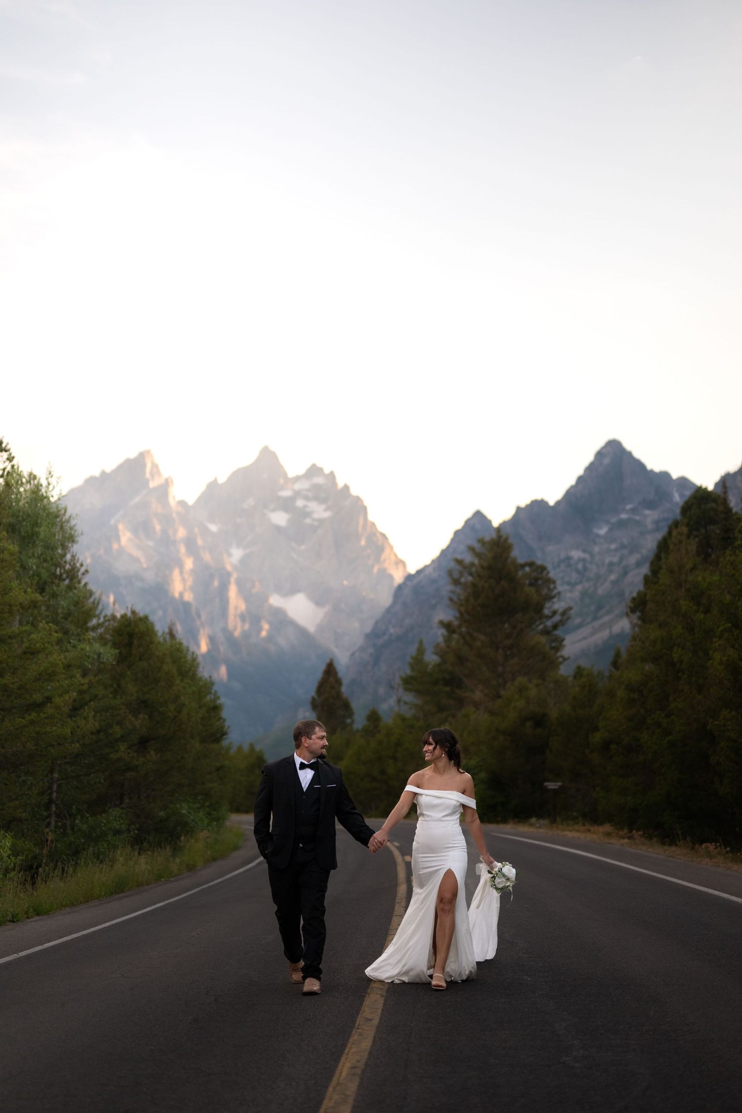 Bride and groom walk hand-in-hand on a mountain road with Grand Teton peaks at sunset.