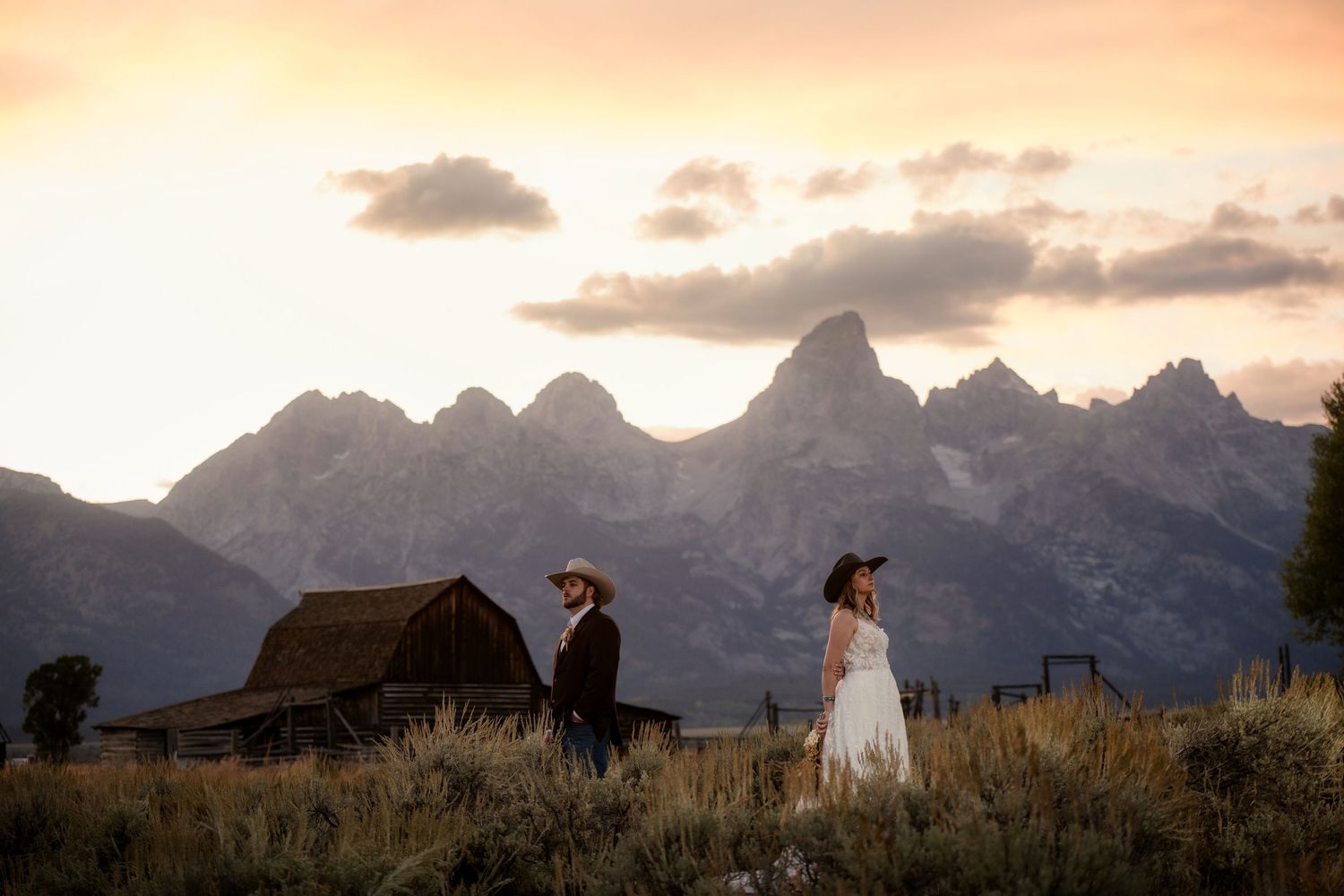 Couple walks through golden fields at sunset with Grand Teton mountains and rustic barn backdrop.