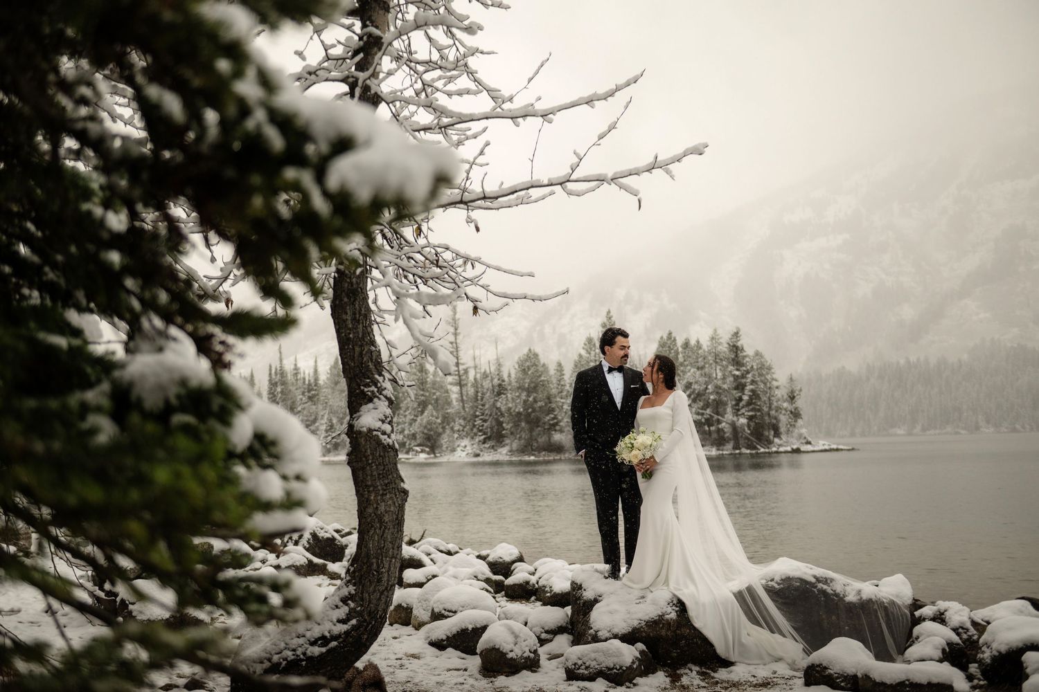 Bride and groom stand by a snowy mountain lake surrounded by snow-dusted pine trees in a winter wedding photo.