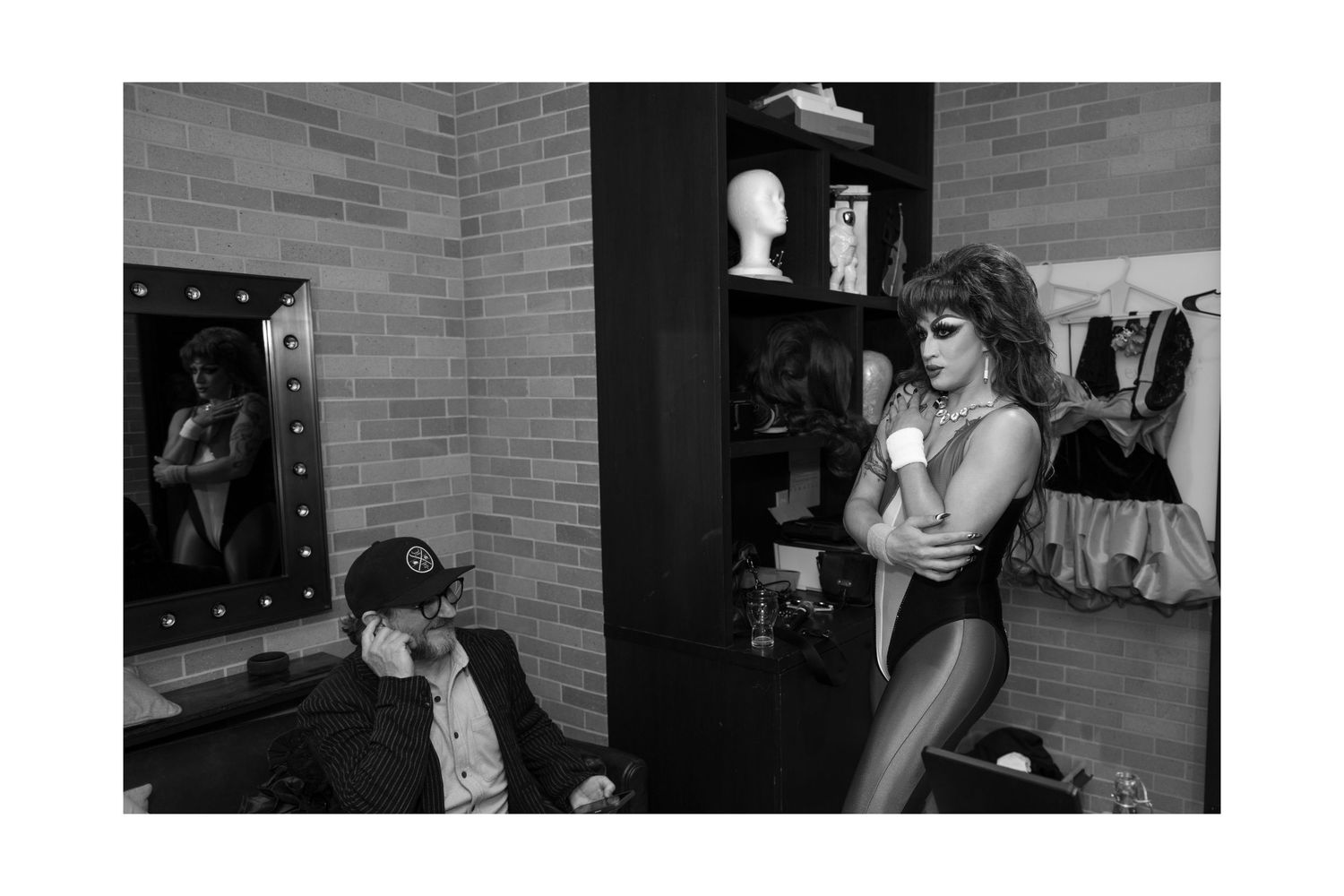 Black and white photo of drag performer in bodysuit with fan in dressing room mirror backstage.
