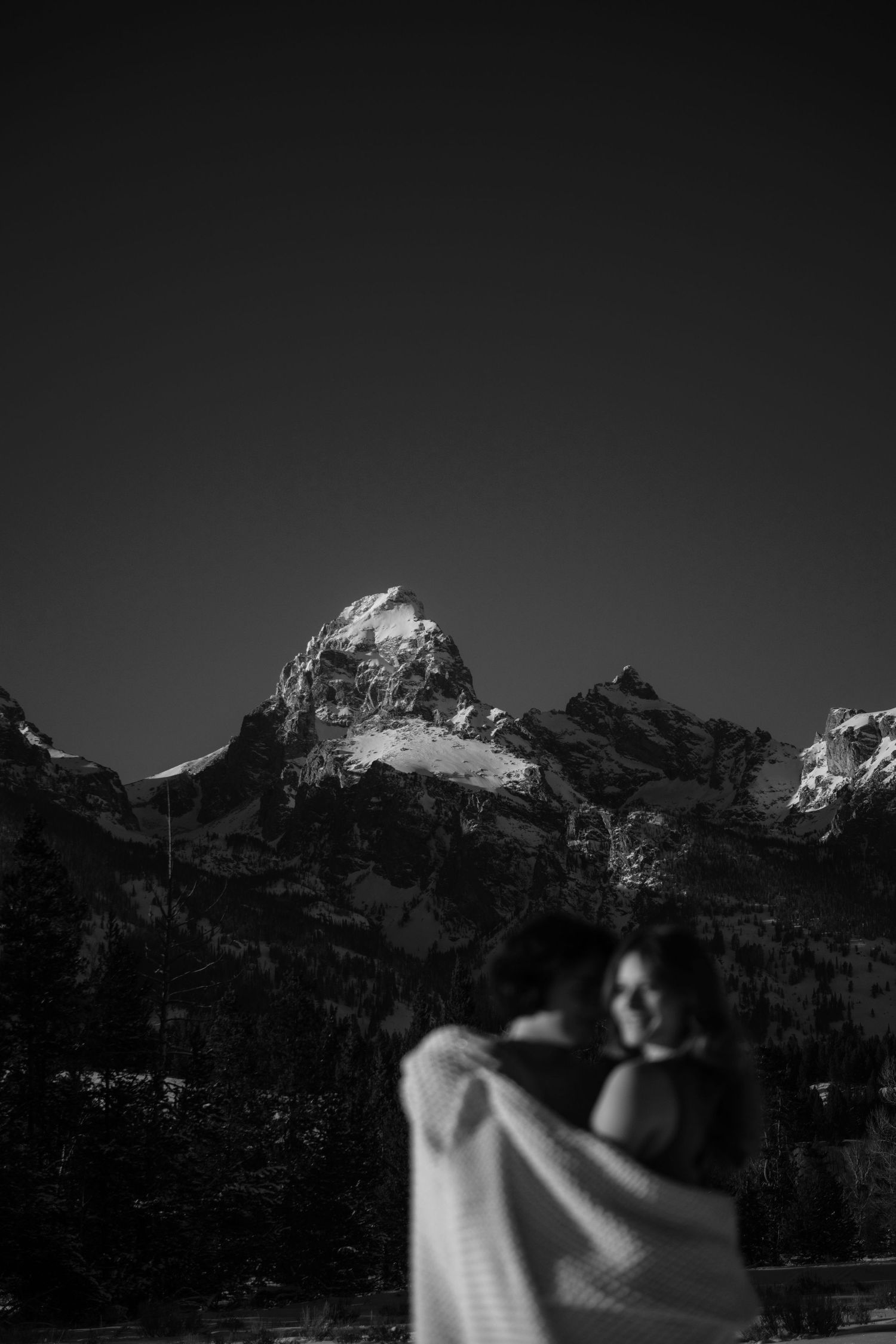 Black and white photo of couple embracing with snow-capped Grand Teton mountains in background.