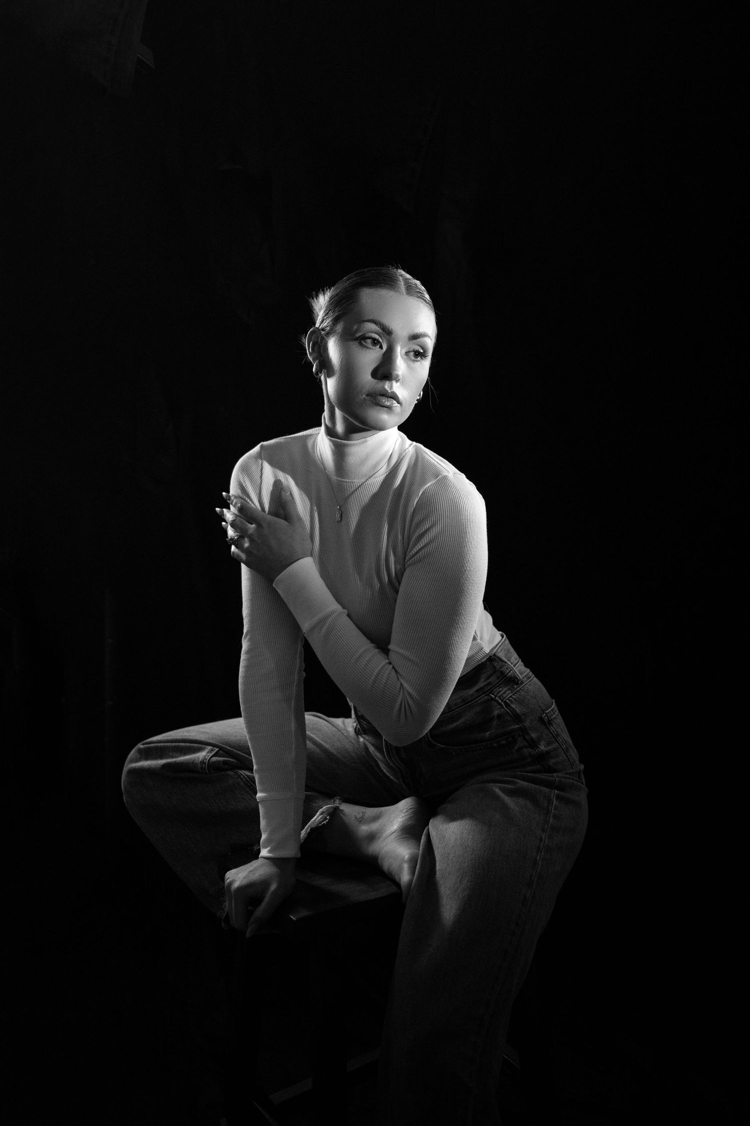 Black and white studio portrait of a young woman in turtleneck and jeans, seated against dark background.