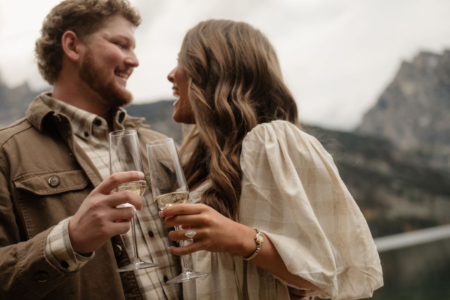 A couple stands together in a mountainous landscape with a lake in the foreground.