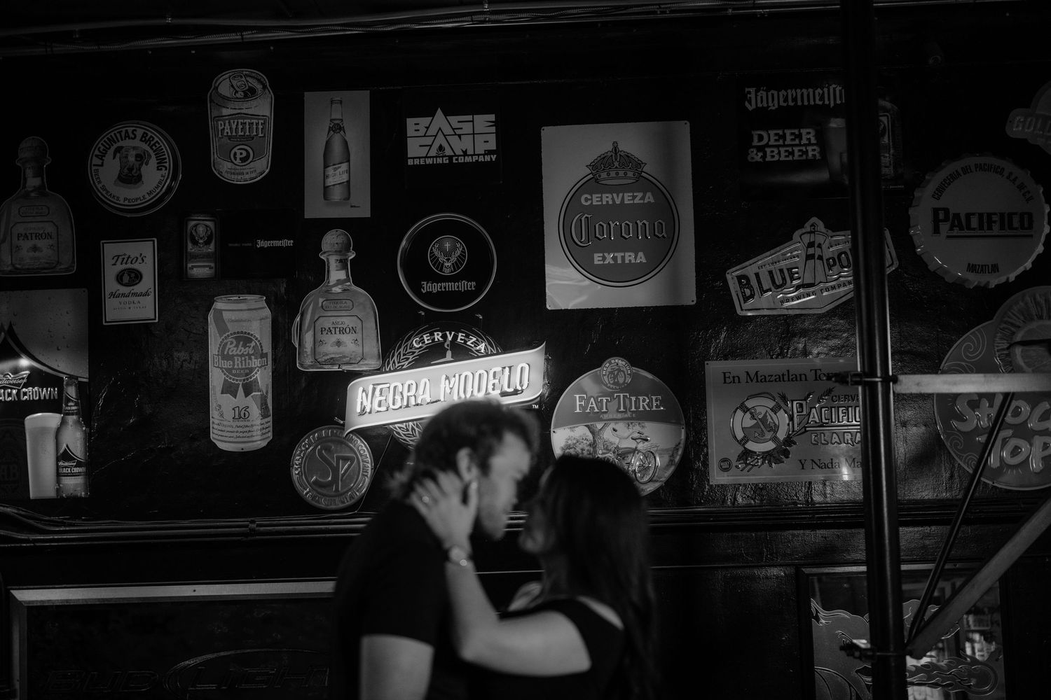 Black and white photo of a couple kissing in a dimly lit bar surrounded by vintage beer signs and neon lights.
