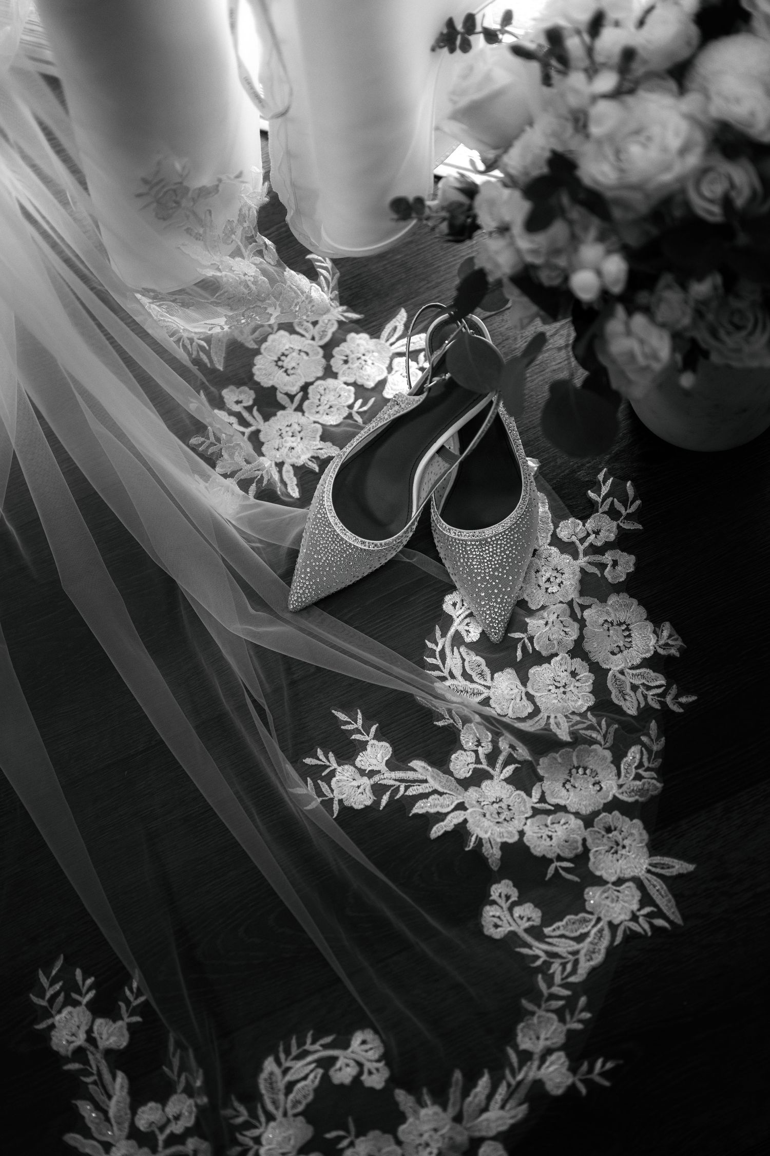 A black and white image of a bridal veil with floral embroidery and a pair of high-heeled shoes.