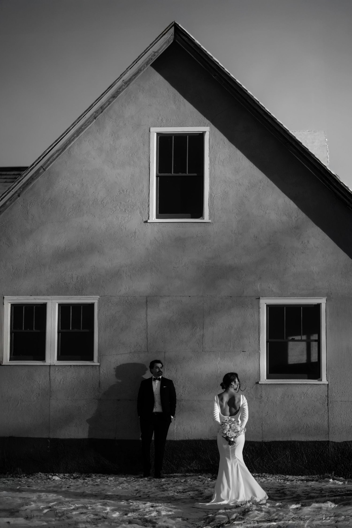 Black and white wedding photo of bride and groom standing apart against a rustic building exterior in winter.