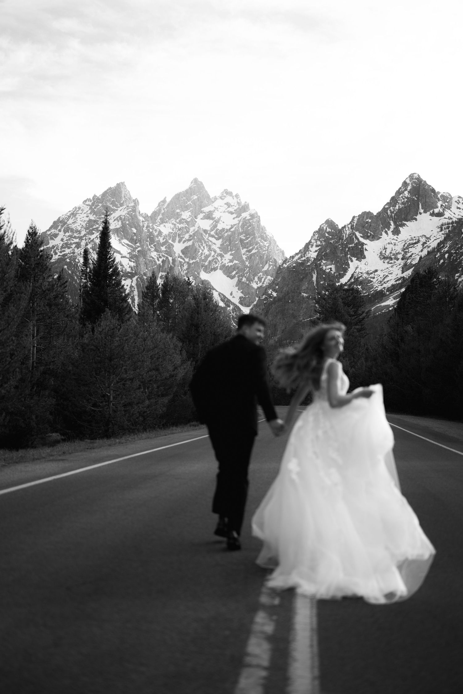 Black and white photo of bride and groom running hand in hand down a road with Grand Teton mountains backdrop.