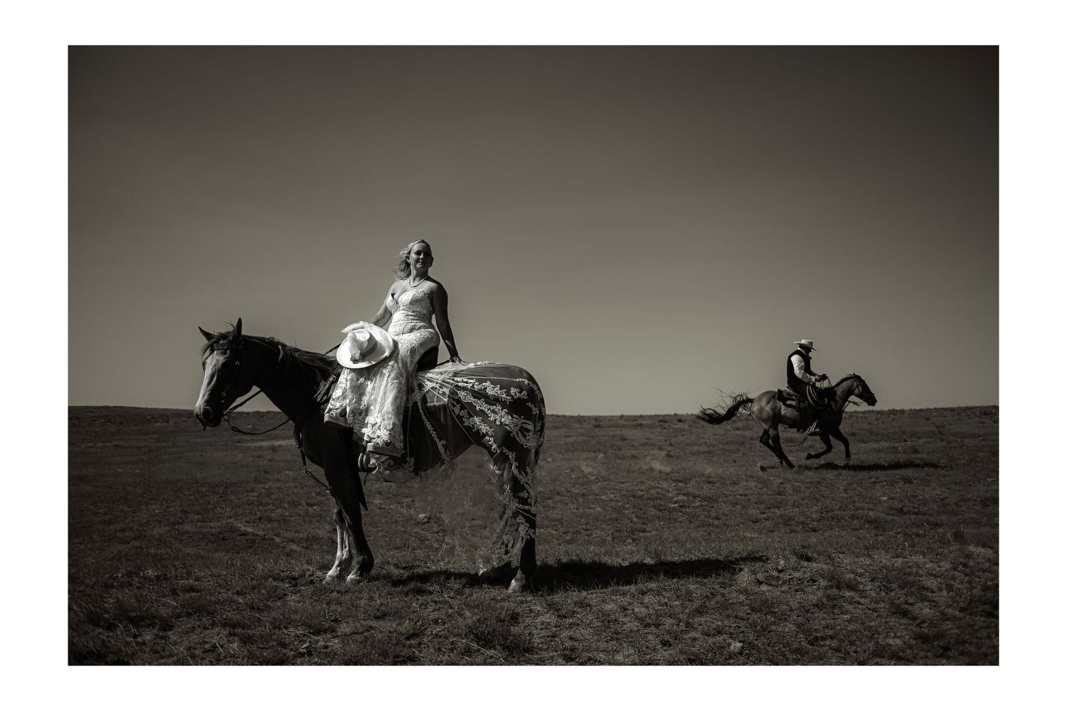 Black and white photo of a Bride in a wedding dress on horseback in an open field with a rider galloping behind. In Jackson Hole Wyoming