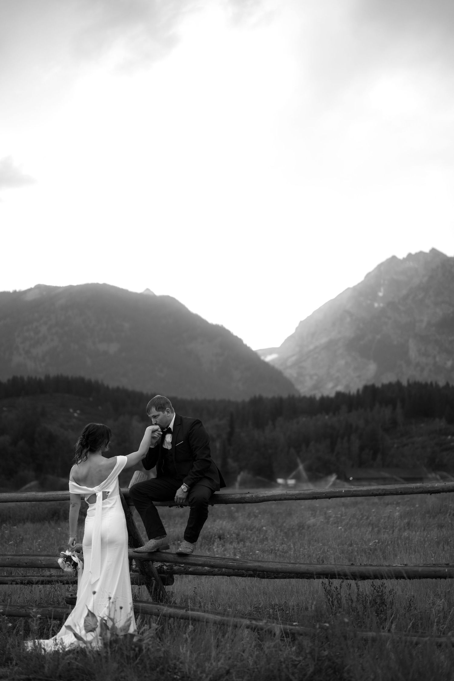 Black and white photo of bride and groom at a rustic fence with mountain backdrop during their wedding day.