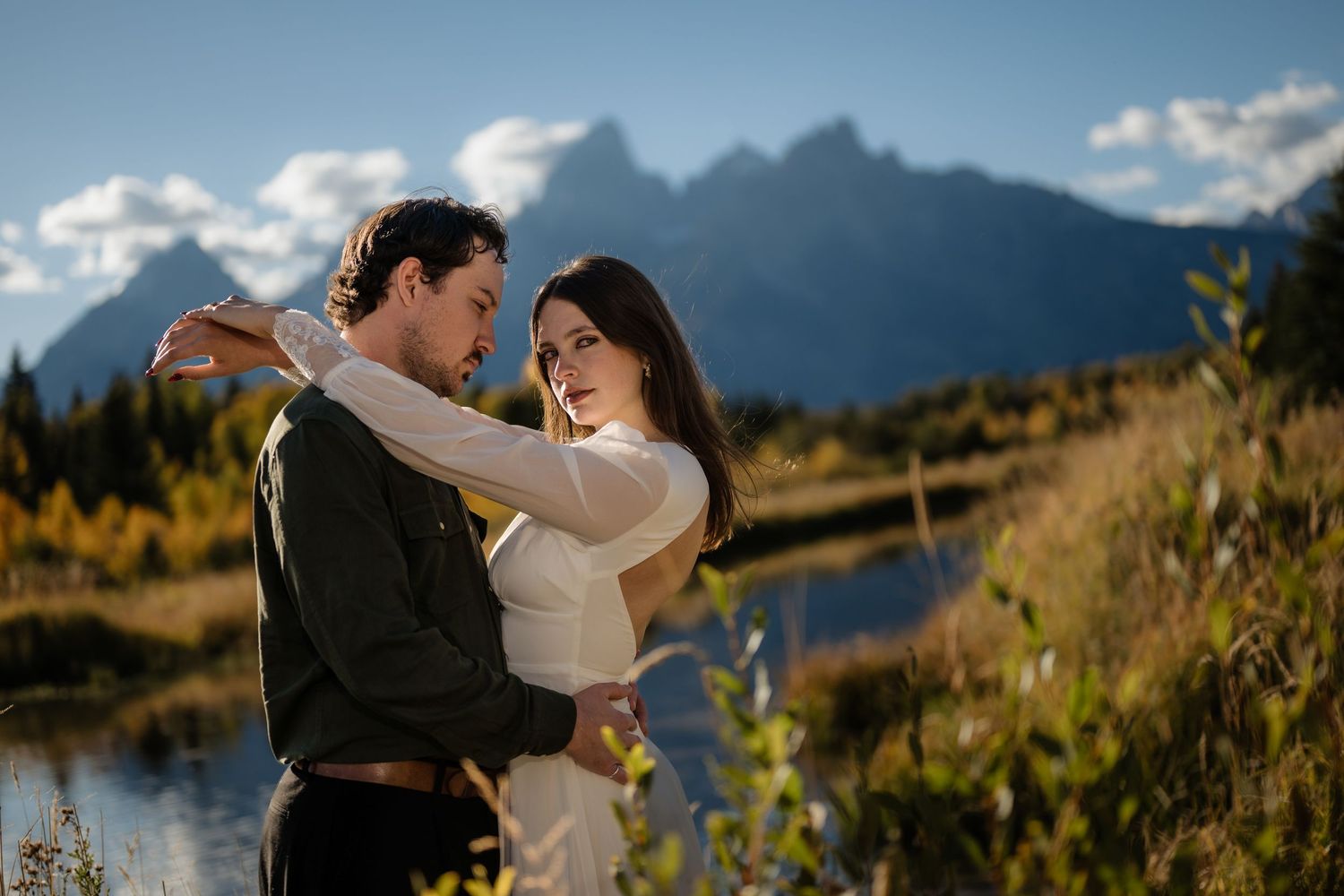 Couple embracing near a mountain lake with Grand Teton peaks and autumn foliage in the background.