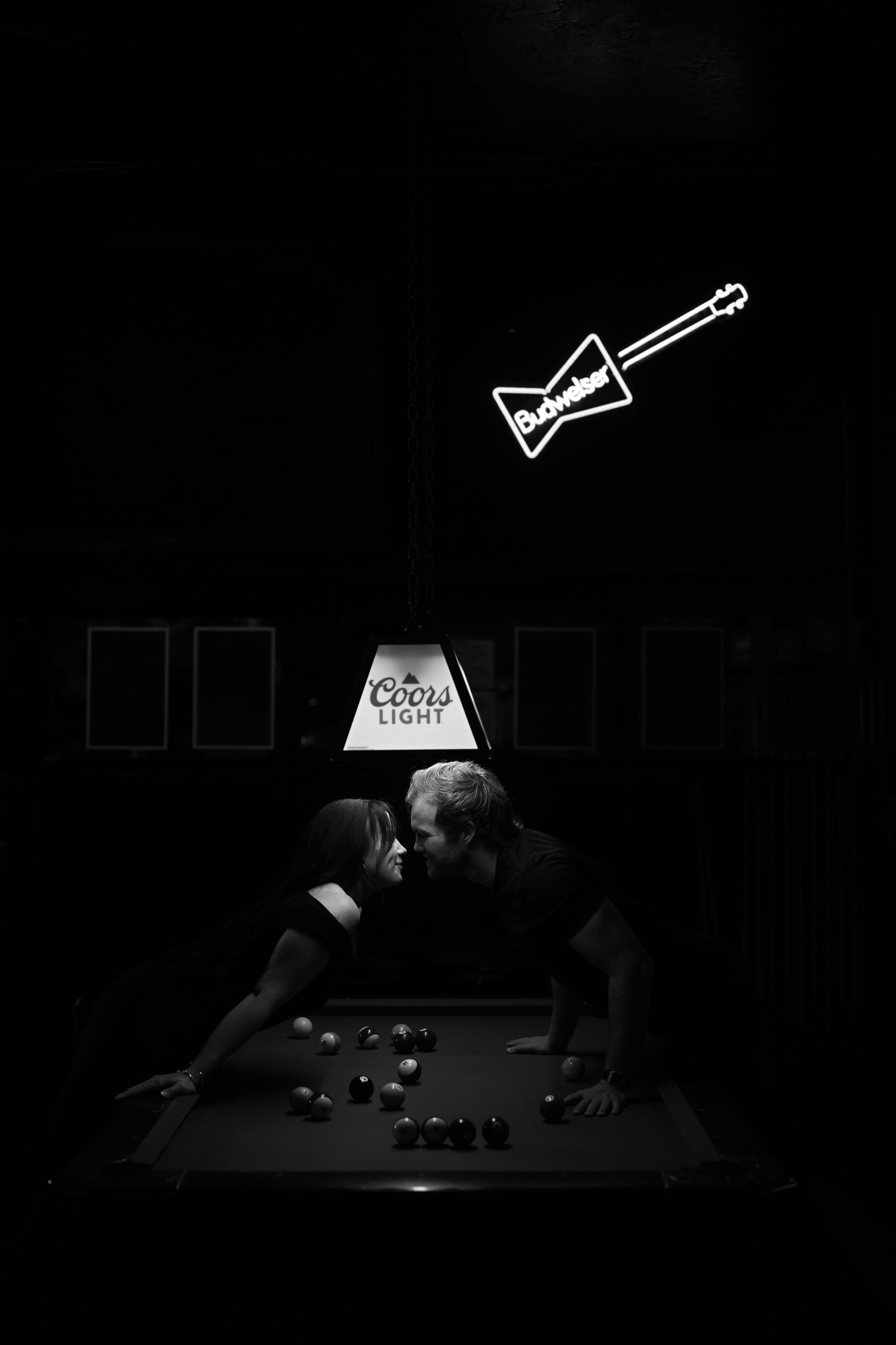 Couple sharing a romantic moment over a pool table under a Coors Light lamp with a Budweiser guitar neon sign above.
