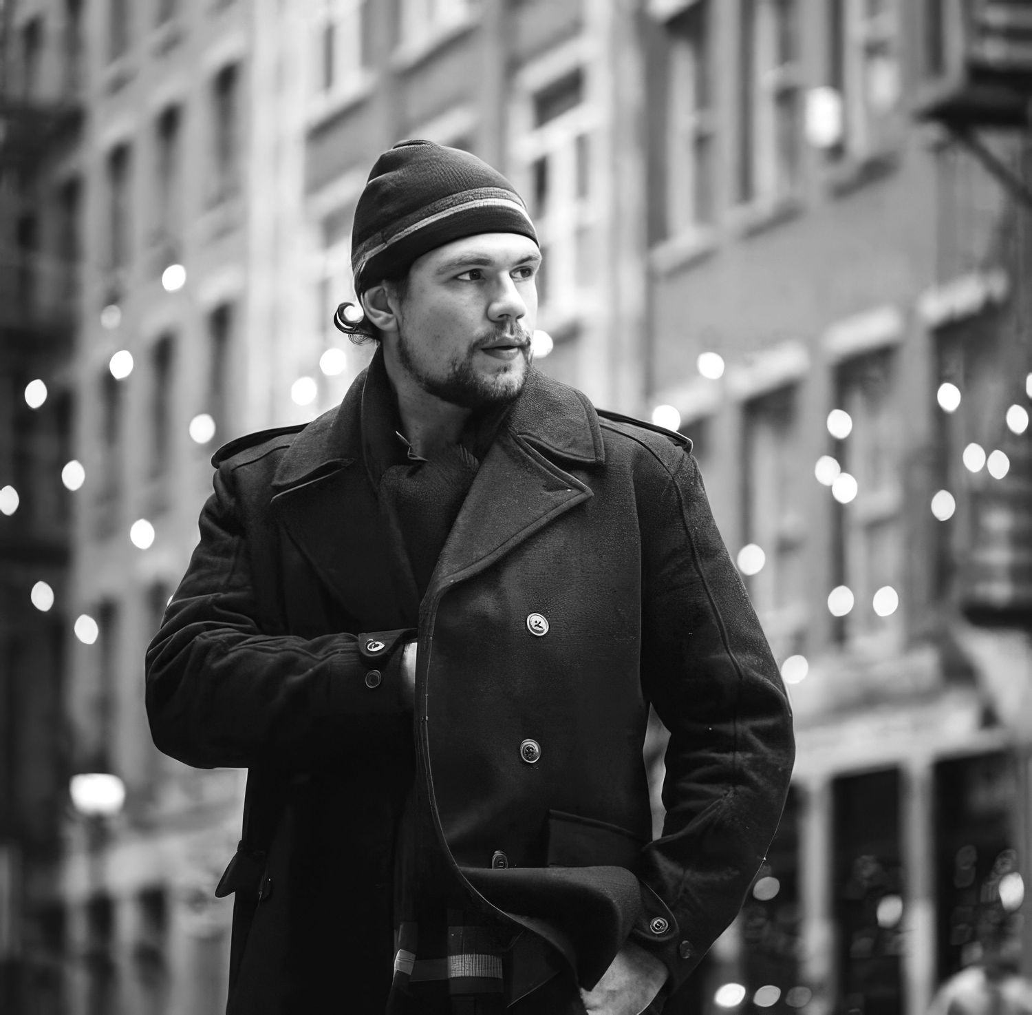 Black and white photo of a man in a peacoat and beanie standing on a city street with bokeh lights.
