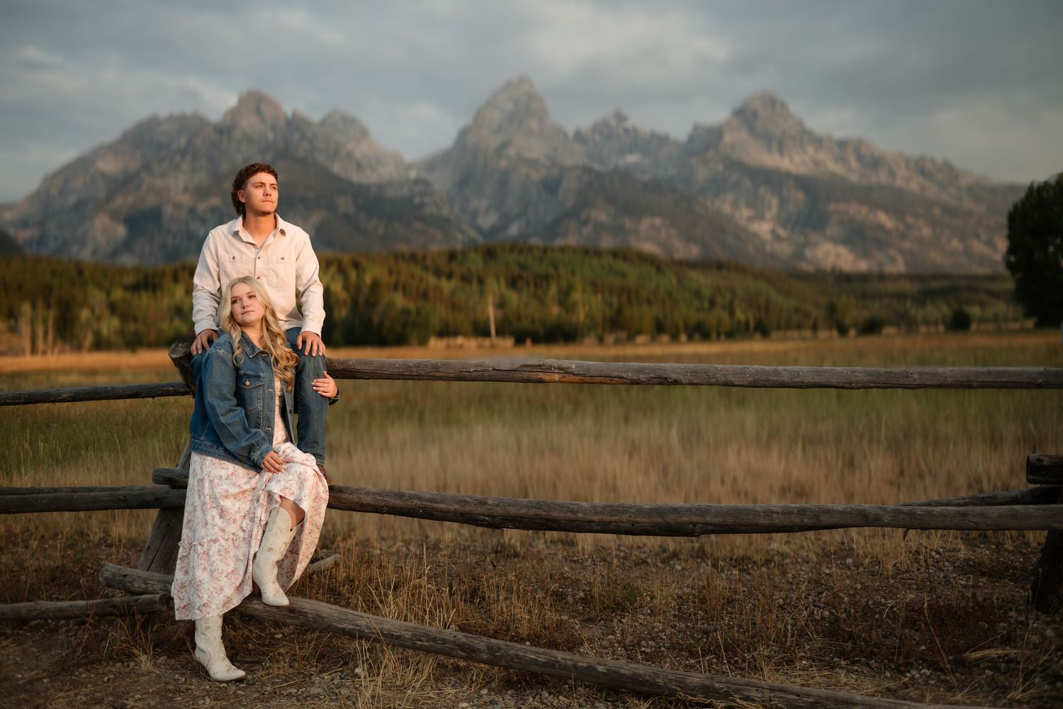 Couple posing by a rustic wooden fence with the Grand Teton mountain range and golden meadows in the background.