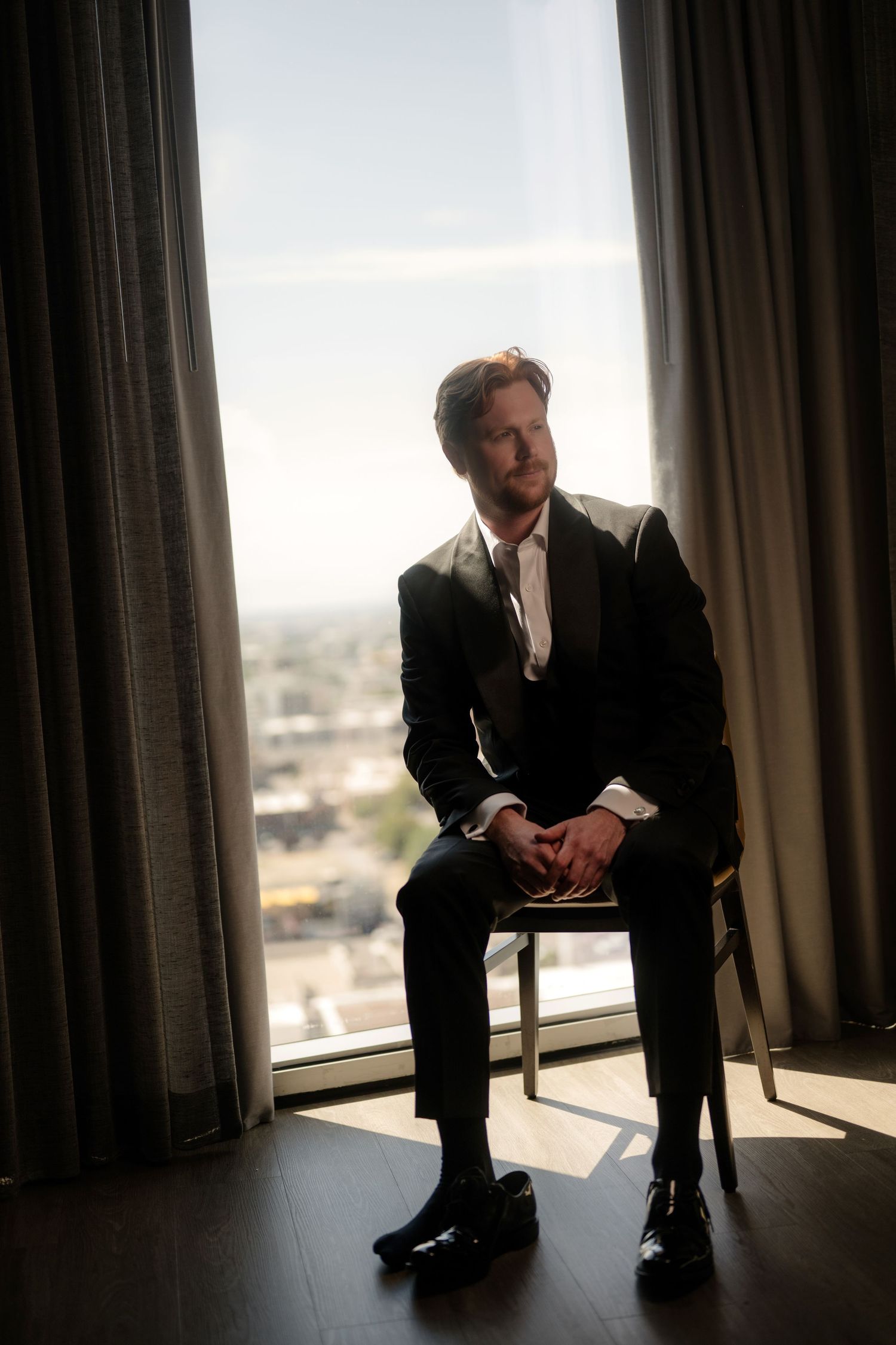 A man in a suit sitting by a window with a city view in the background.
