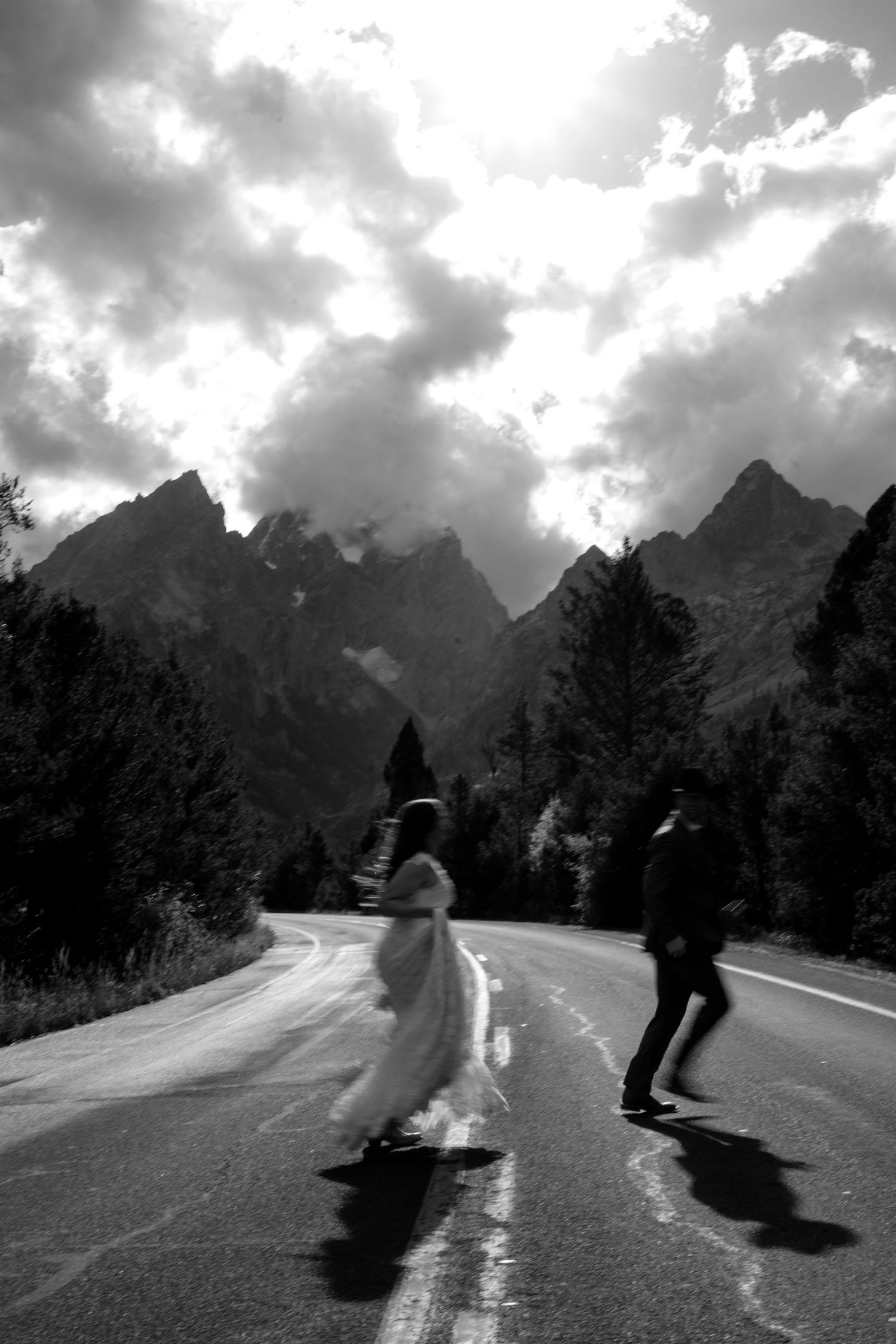 Black and white photo of bride and groom crossing a mountain road with dramatic clouds and peaks in the background.