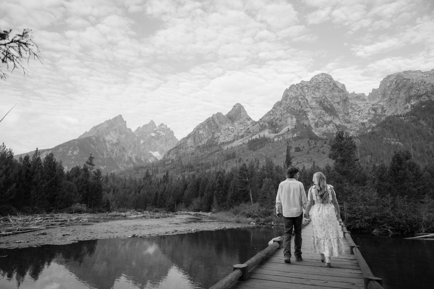 A black and white image of a couple walking on a wooden dock by a lake with mountains in the background.
