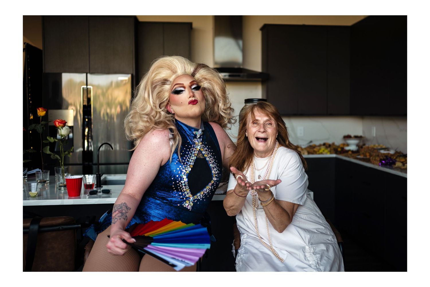 A drag queen in colorful outfit poses with an elderly woman in a kitchen setting.