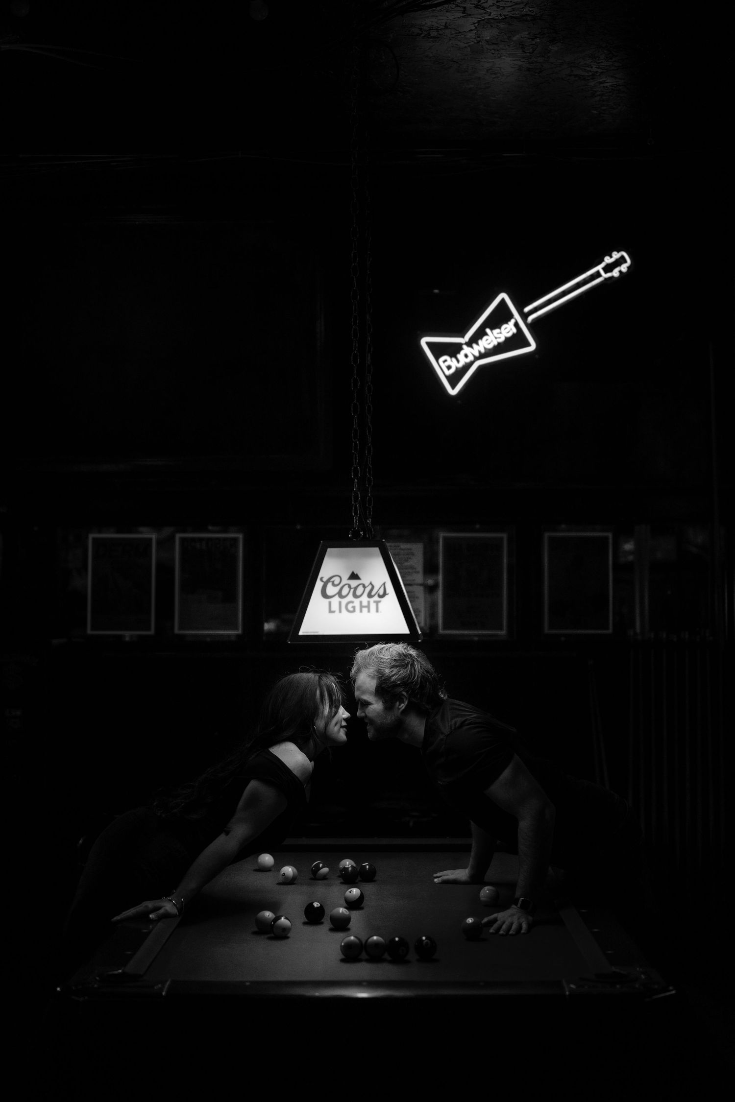 Couple sharing a kiss over a pool table under Coors Light lamp with Budweiser guitar neon sign in dark bar.