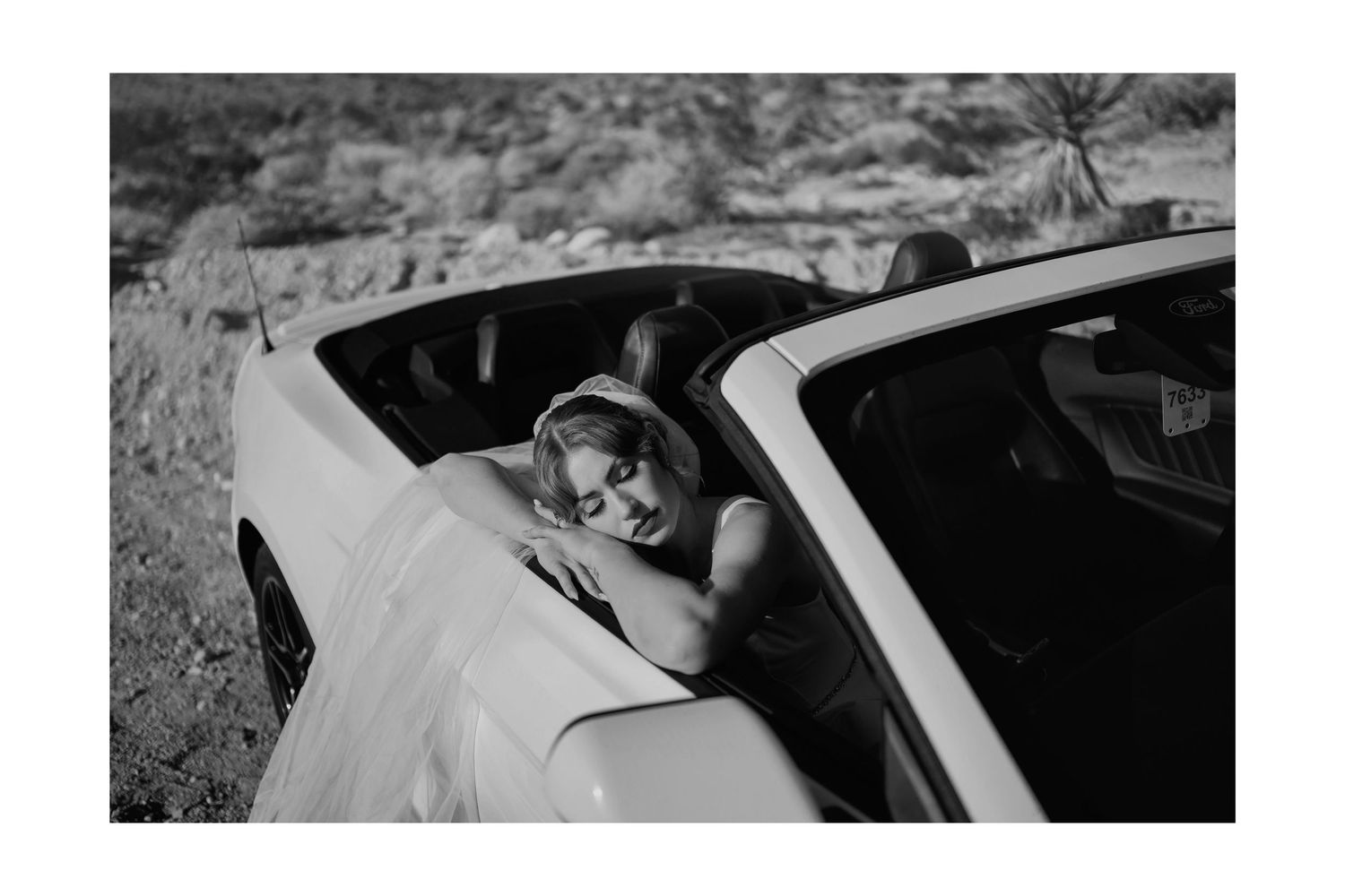 Black and white photo of a bride in a wedding dress leaning on a convertible car in a desert landscape.