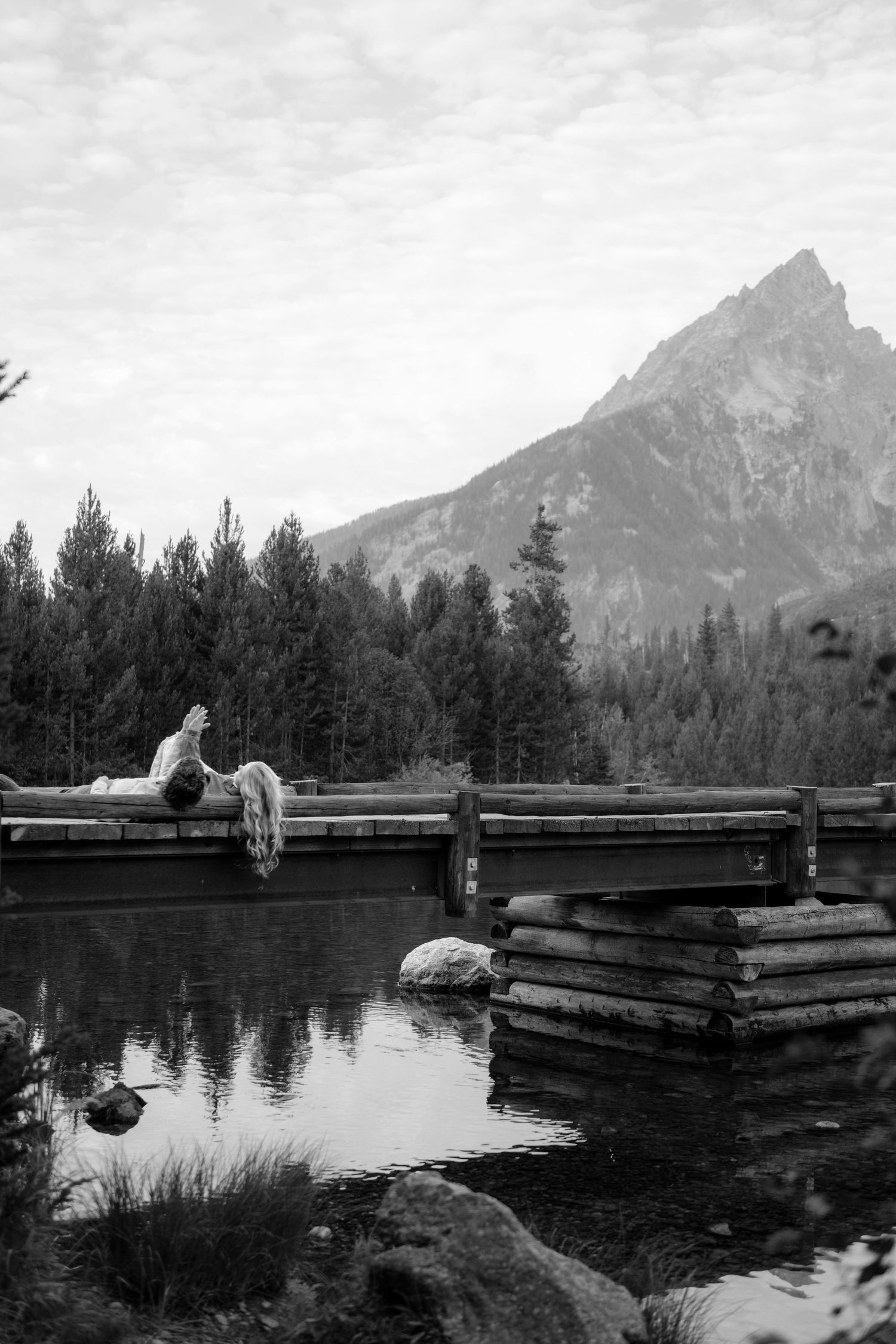 Black and white photo of person relaxing on wooden bridge over calm mountain lake with pine trees and peak backdrop.