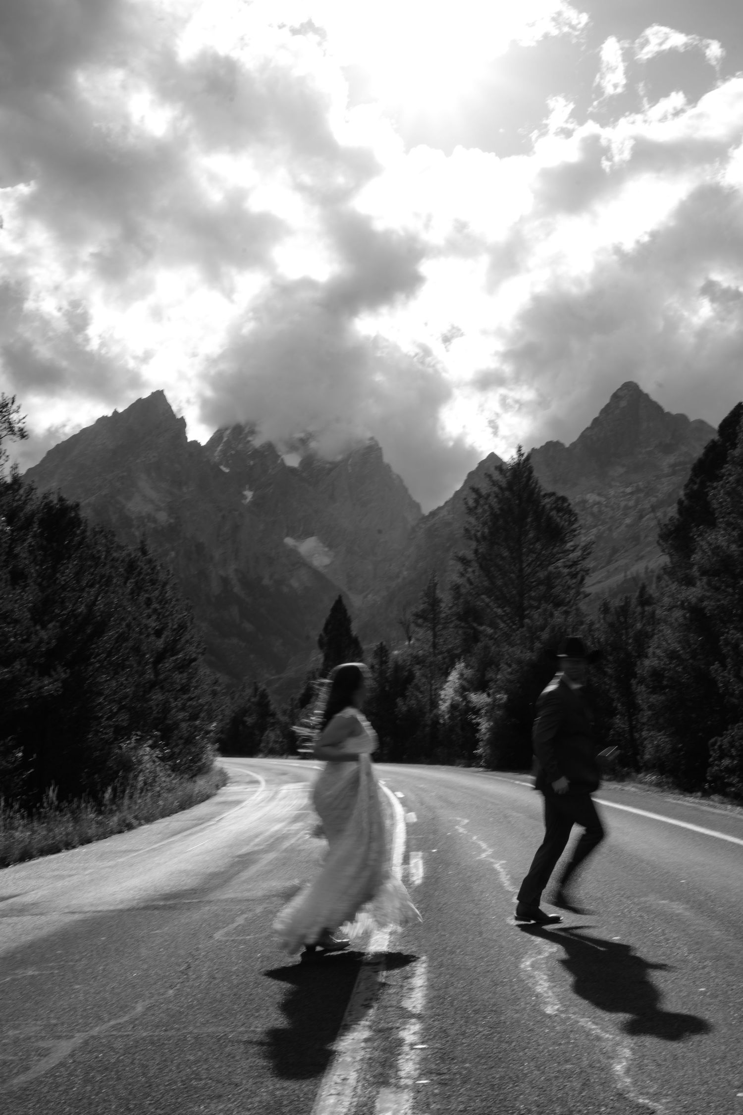 Black and white photo of a bride and groom crossing a mountain road with dramatic clouds and peaks in the background.