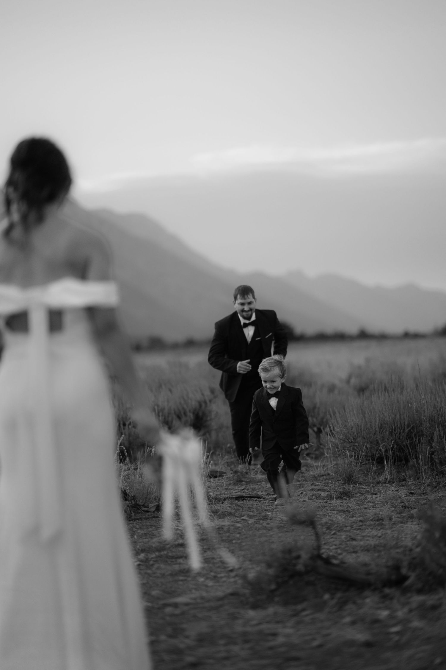 A woman in a white dress stands in a field, with a man in a suit walking towards her in the distance.