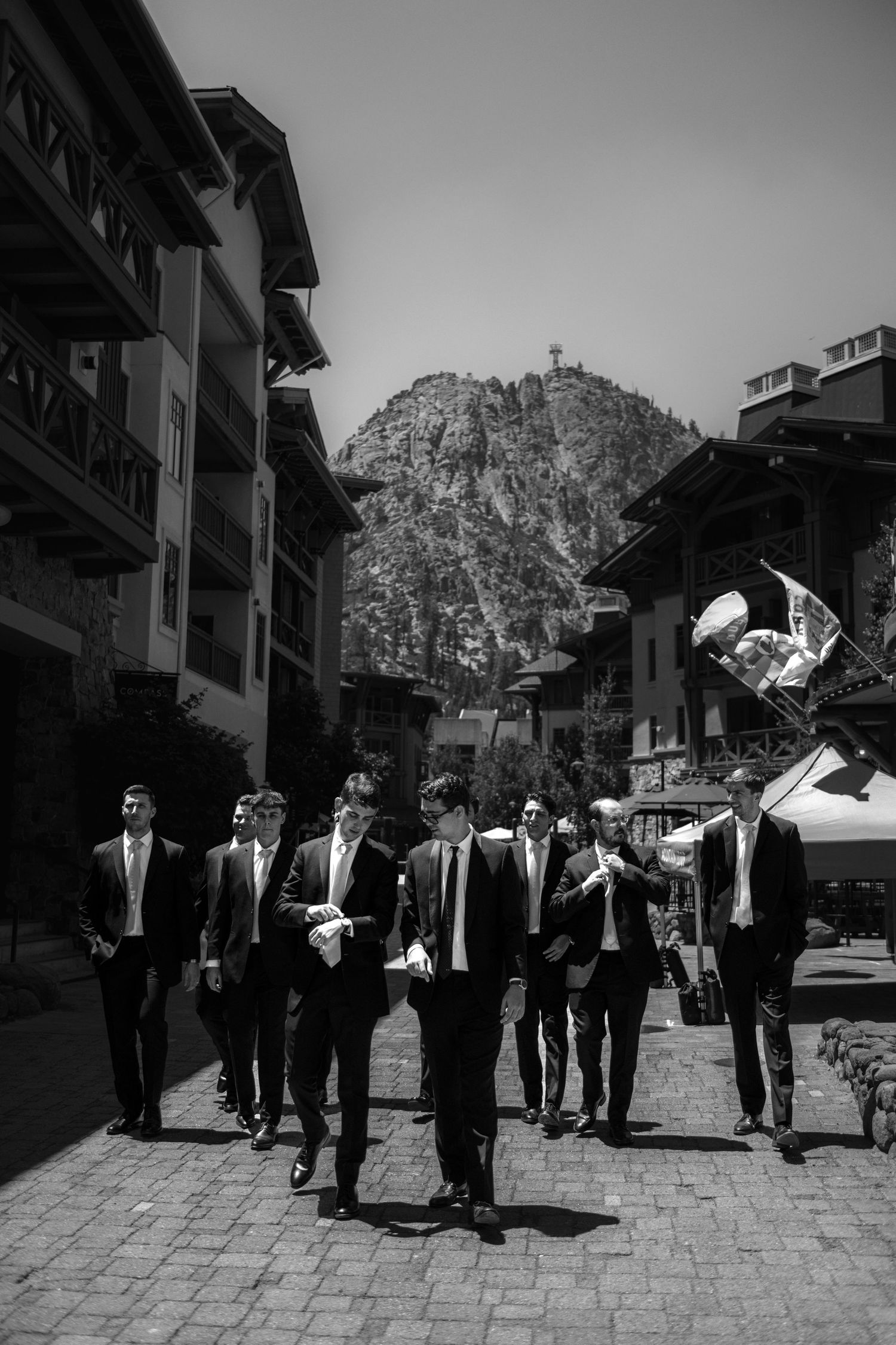 A black and white image of a group of people walking through a narrow street with buildings and a large mountain in the background.