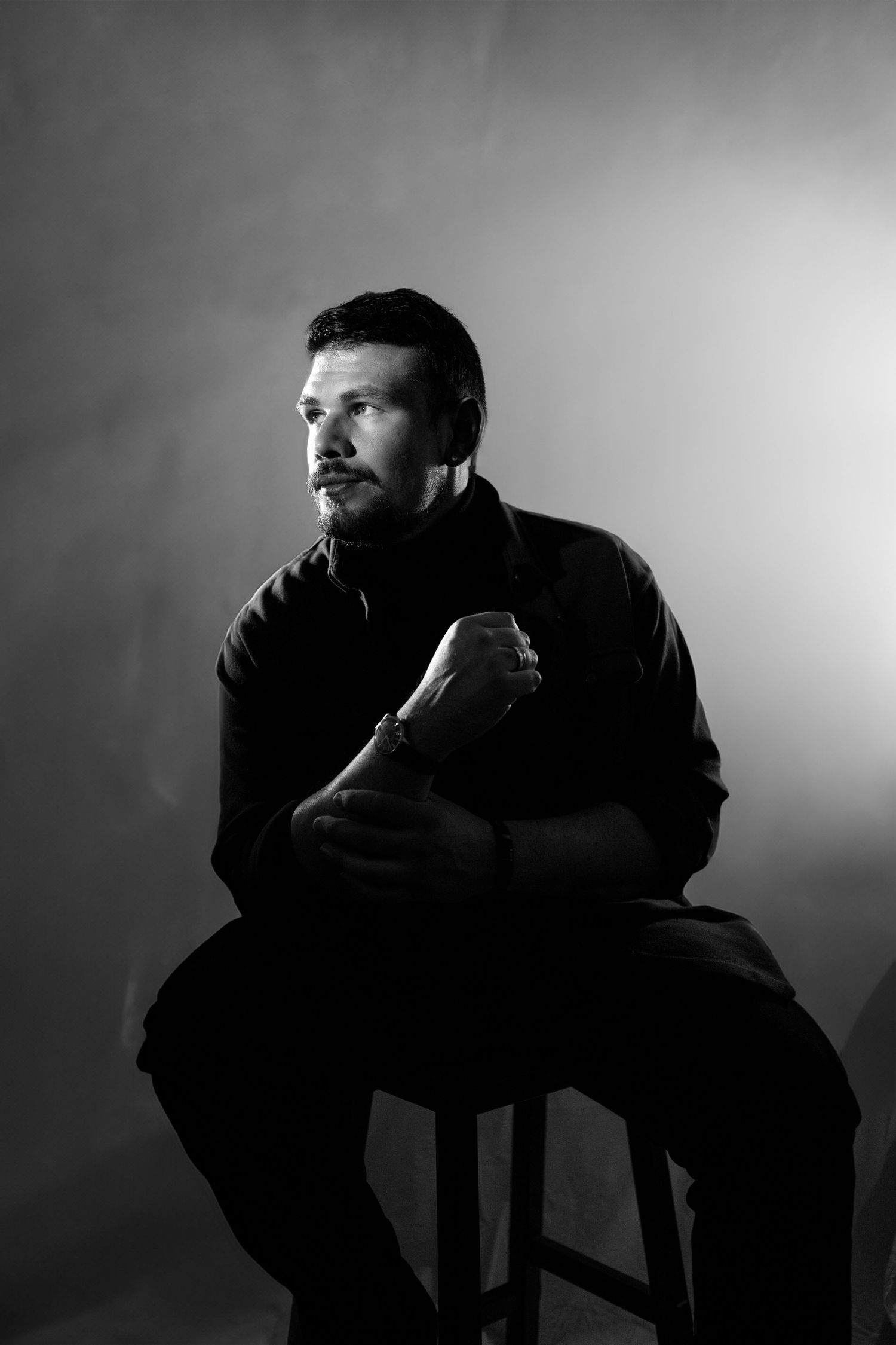 Black and white portrait of a bearded man sitting on a stool, arms crossed, gazing thoughtfully in dramatic studio lighting.