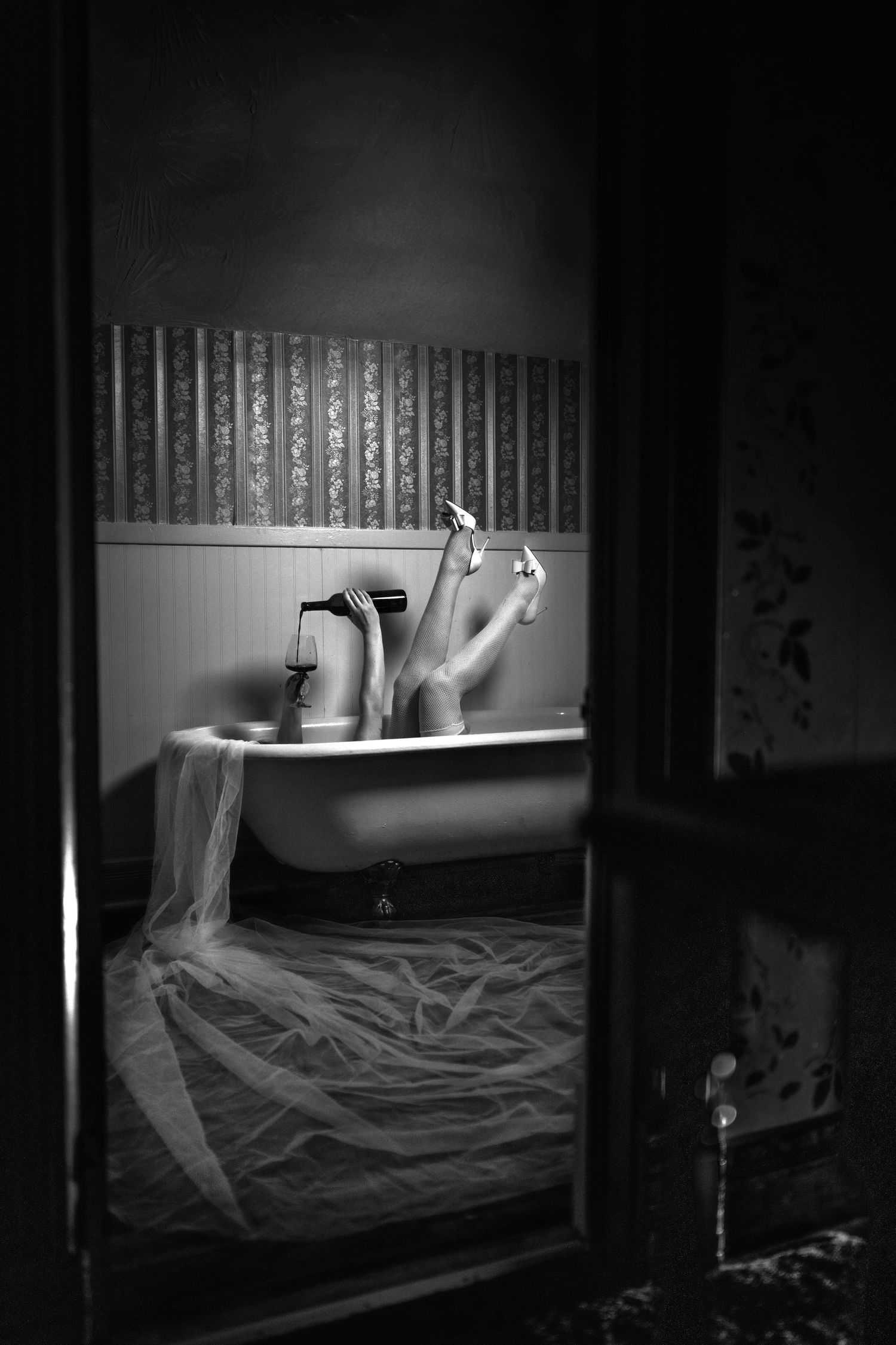 Black and white photo of a person relaxing in a vintage clawfoot bathtub with sheer fabric draped over the side.